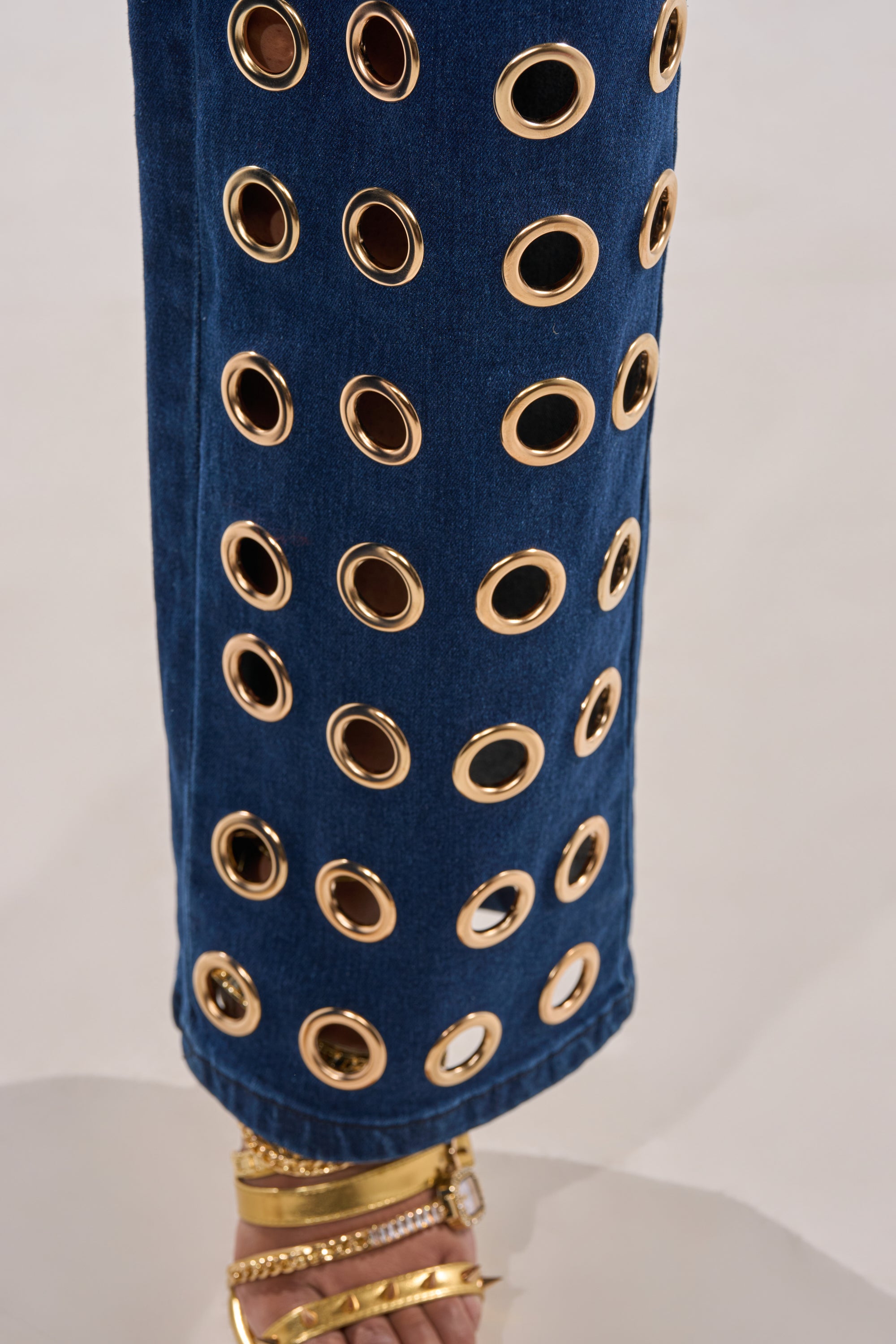 Close-up of SEE THROUGH ME WIDE LEG DENIM, high-waisted with multiple rows of large gold grommets. The straight-leg jeans end above the ankle, revealing sandal gold straps for a bold statement look. 6