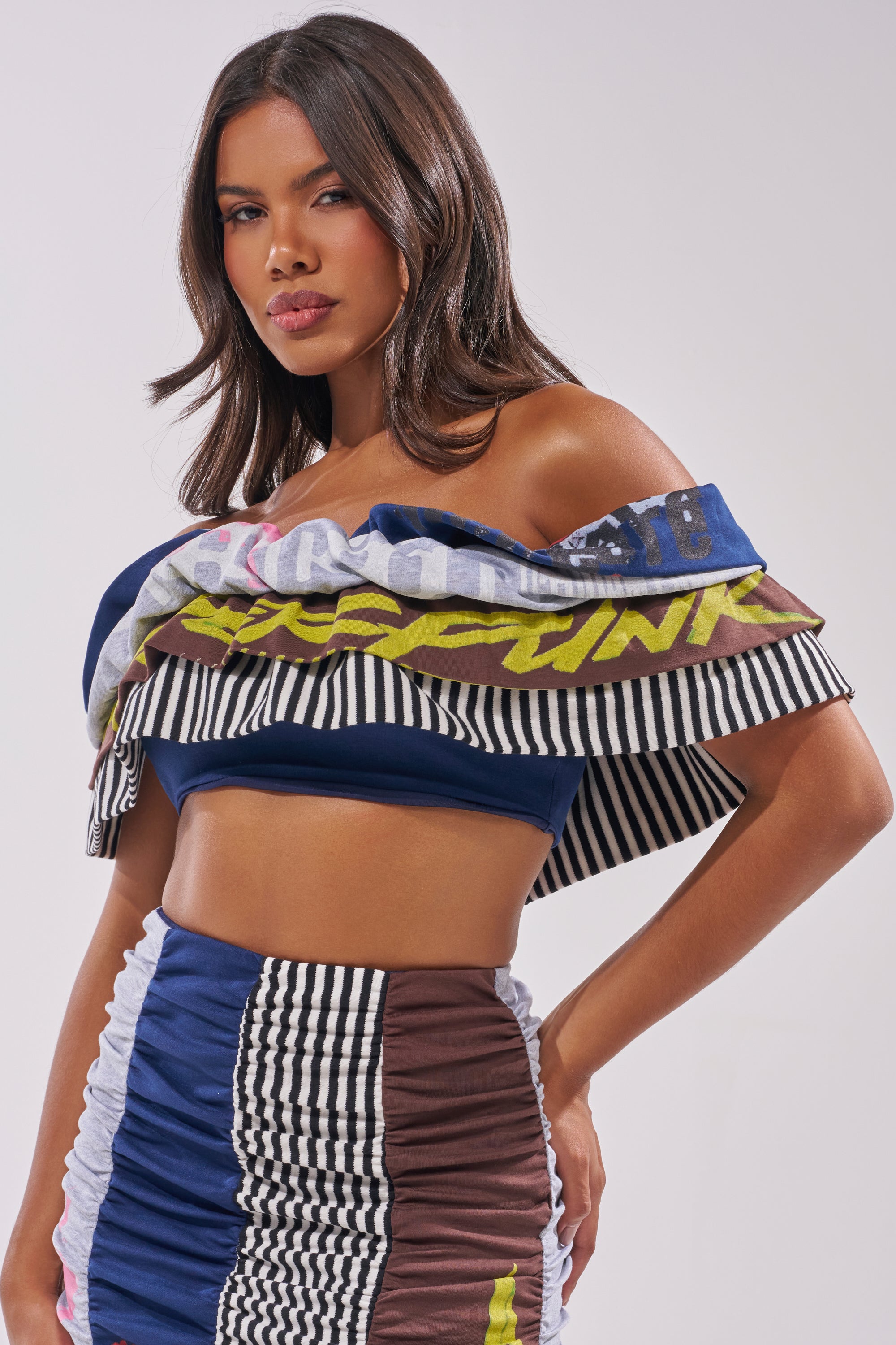 A woman with medium-length brown hair and a neutral expression poses confidently in the THAT GIRL GRAPHIC TEE TOP, an off-shoulder crop top with striped and graphic patterns, paired with a matching high-waisted skirt. 2