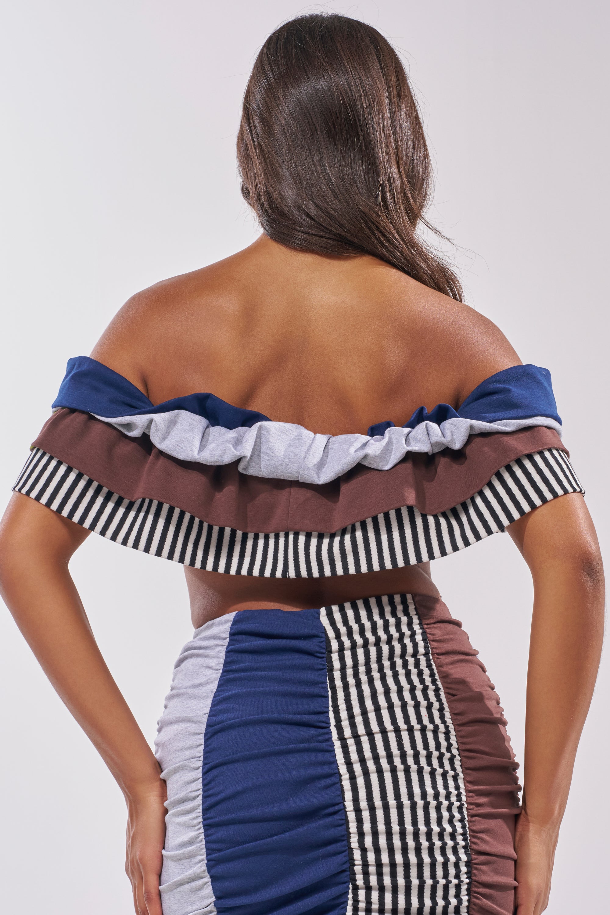 A woman stands with her back to the camera in the THAT GIRL GRAPHIC TEE TOP, styled upcycled with blue, brown, gray, and black-and-white striped panels. Her long hair flows loose. 4