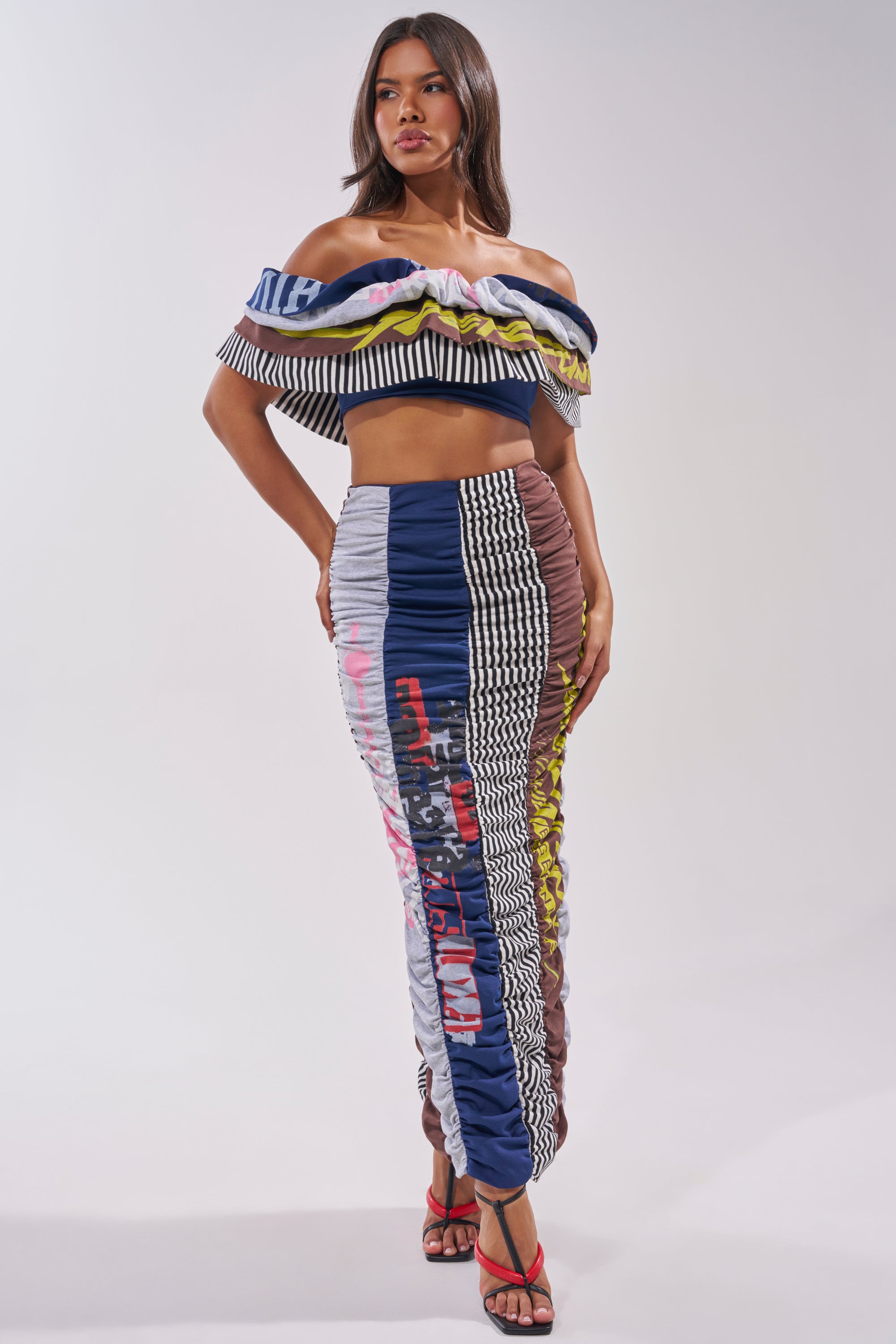 A woman models the THAT GIRL GRAPHIC TEE MAXI SKIRT, featuring mixed patterns and text. She pairs it with a colorful crop top and strappy heels, posing against a plain white background. 0