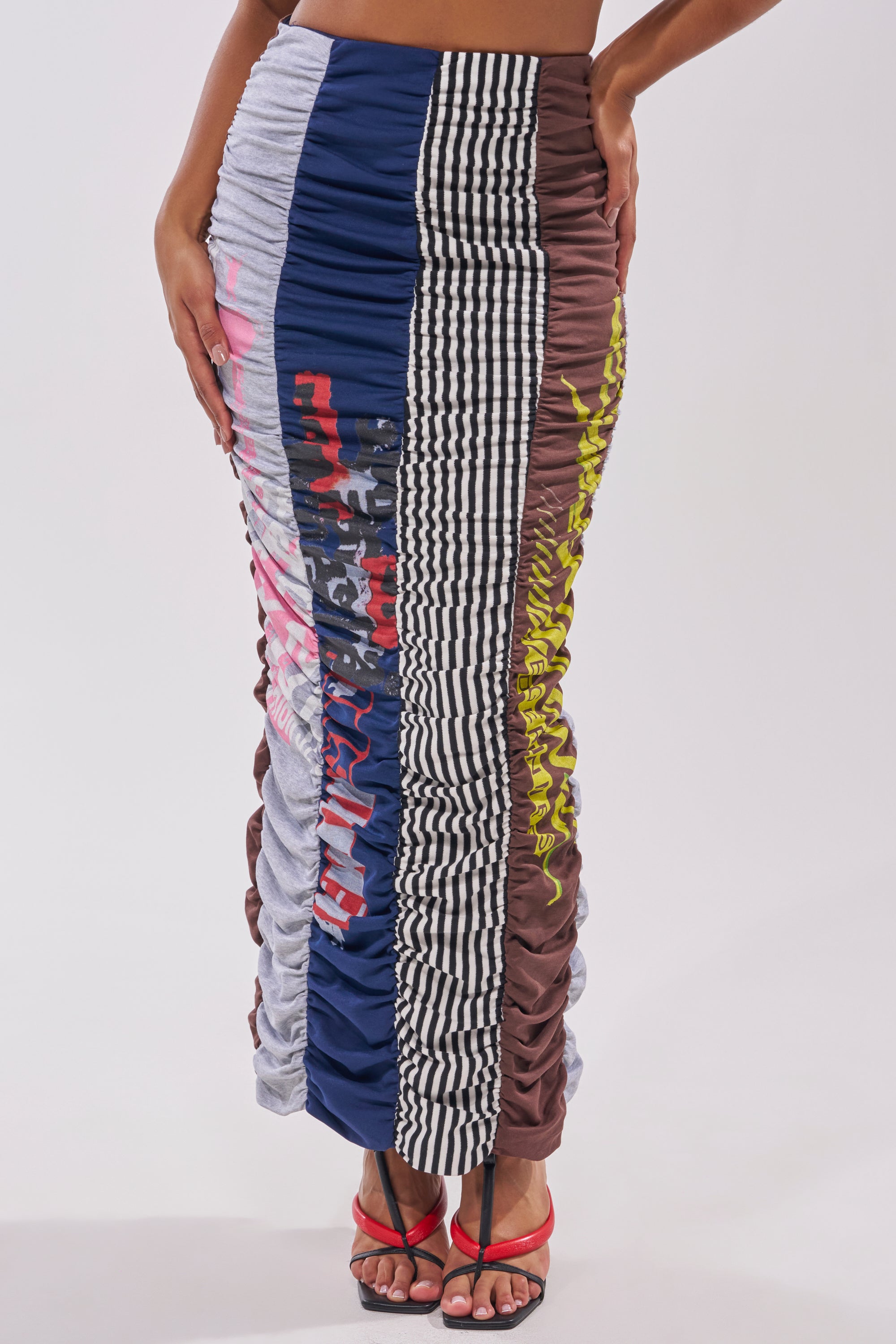 Wearing the THAT GIRL GRAPHIC TEE MAXI SKIRT, a long patchwork skirt with blue, grey, brown, and black-and-white check panels plus graphic prints, styled with black and red strappy heels. 1