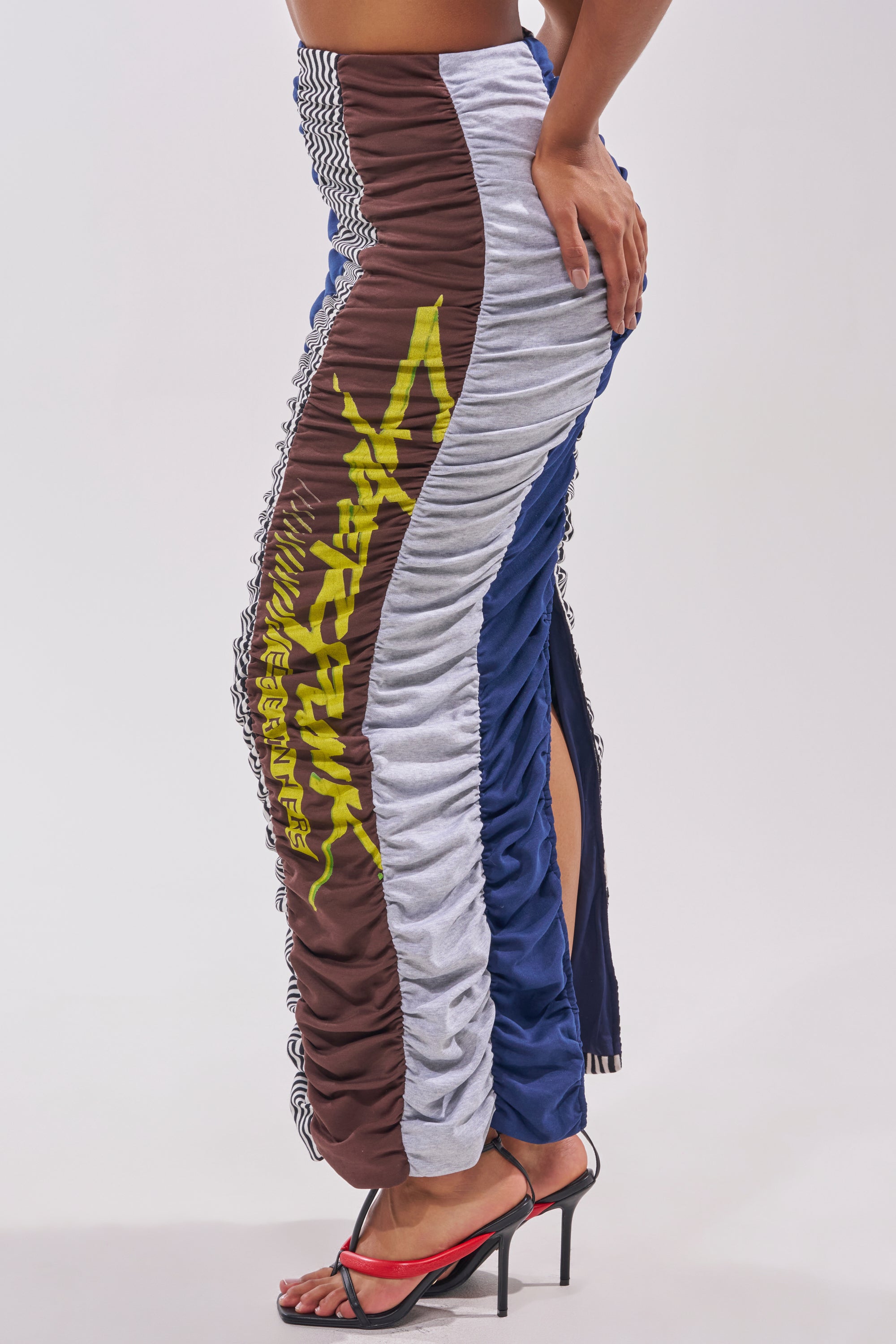 Someone styling the THAT GIRL GRAPHIC TEE MAXI SKIRT—an upcycled-inspired, color-blocked maxi with brown, blue, and gray panels, yellow graphic text, and a side slit—teamed with red high-heeled sandals. 2