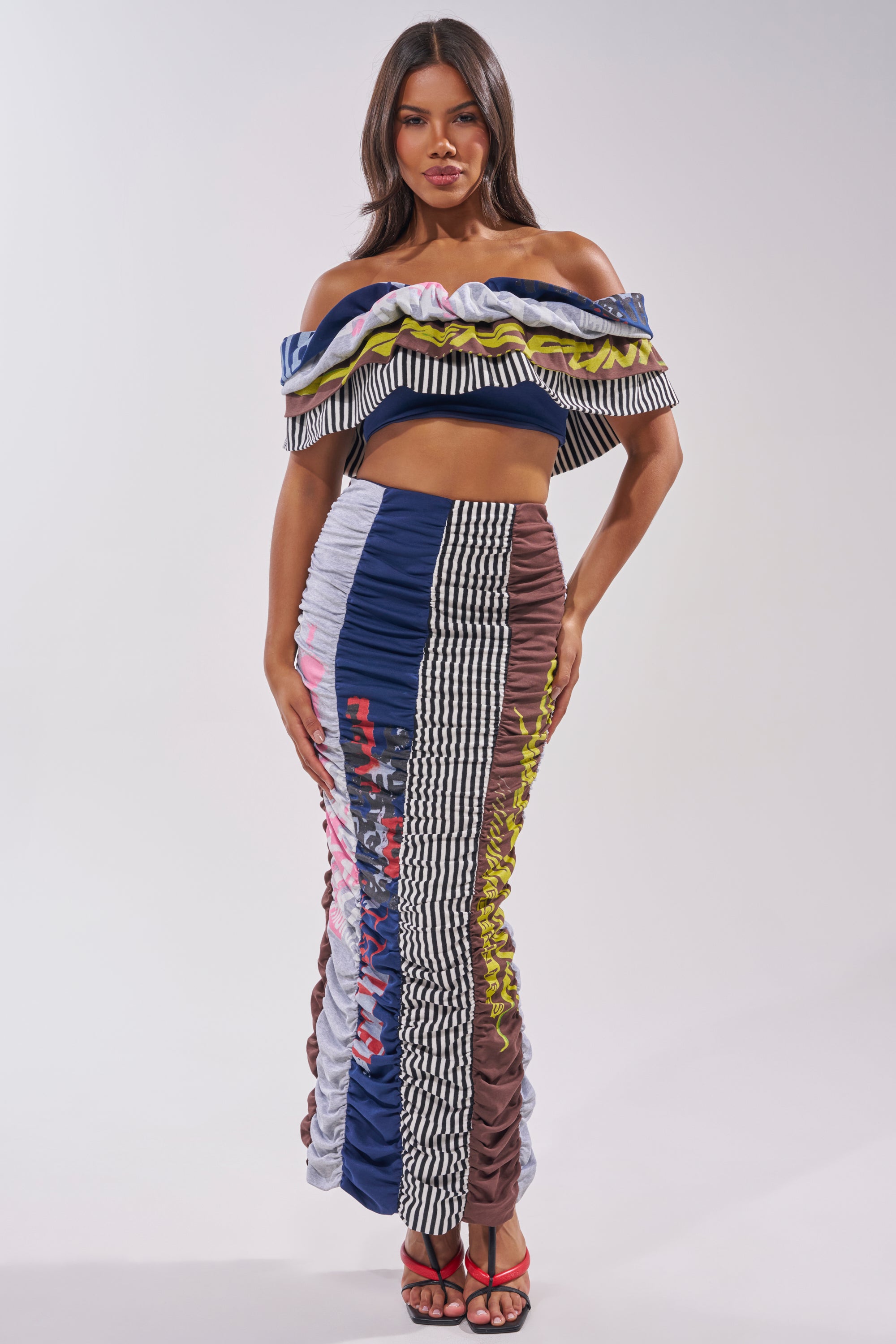 A woman wears the THAT GIRL GRAPHIC TEE MAXI SKIRT, a colorful patchwork maxi skirt with vertical stripes and patterns in blue, yellow, black, white, and brown. She pairs it with a graphic tee top and black heels. 5