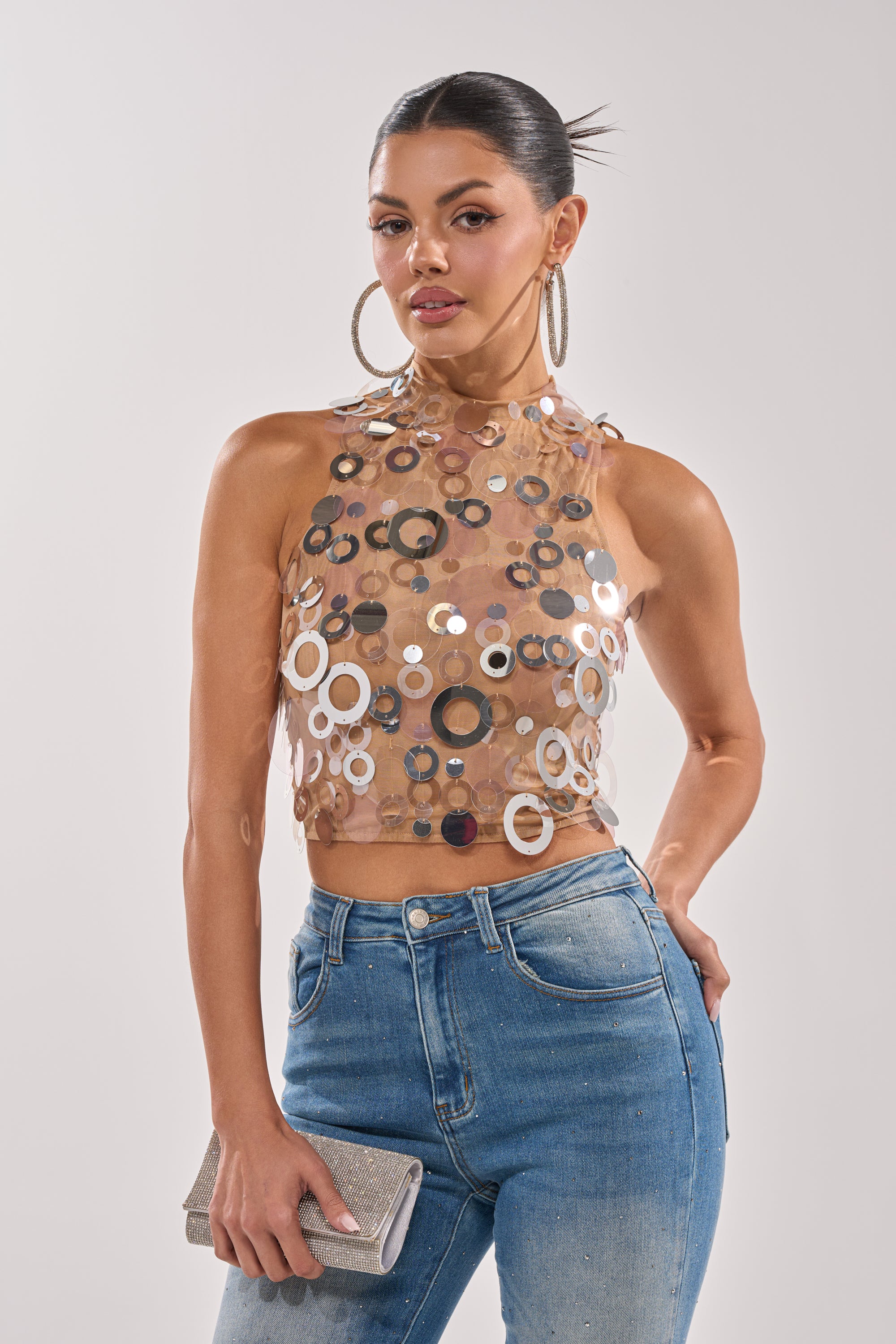 A woman with slicked-back hair wears the BLING BLING TANK—a sequin-embellished party top—paired with blue jeans and large hoop earrings, holding a glittery clutch against a plain light background. 0