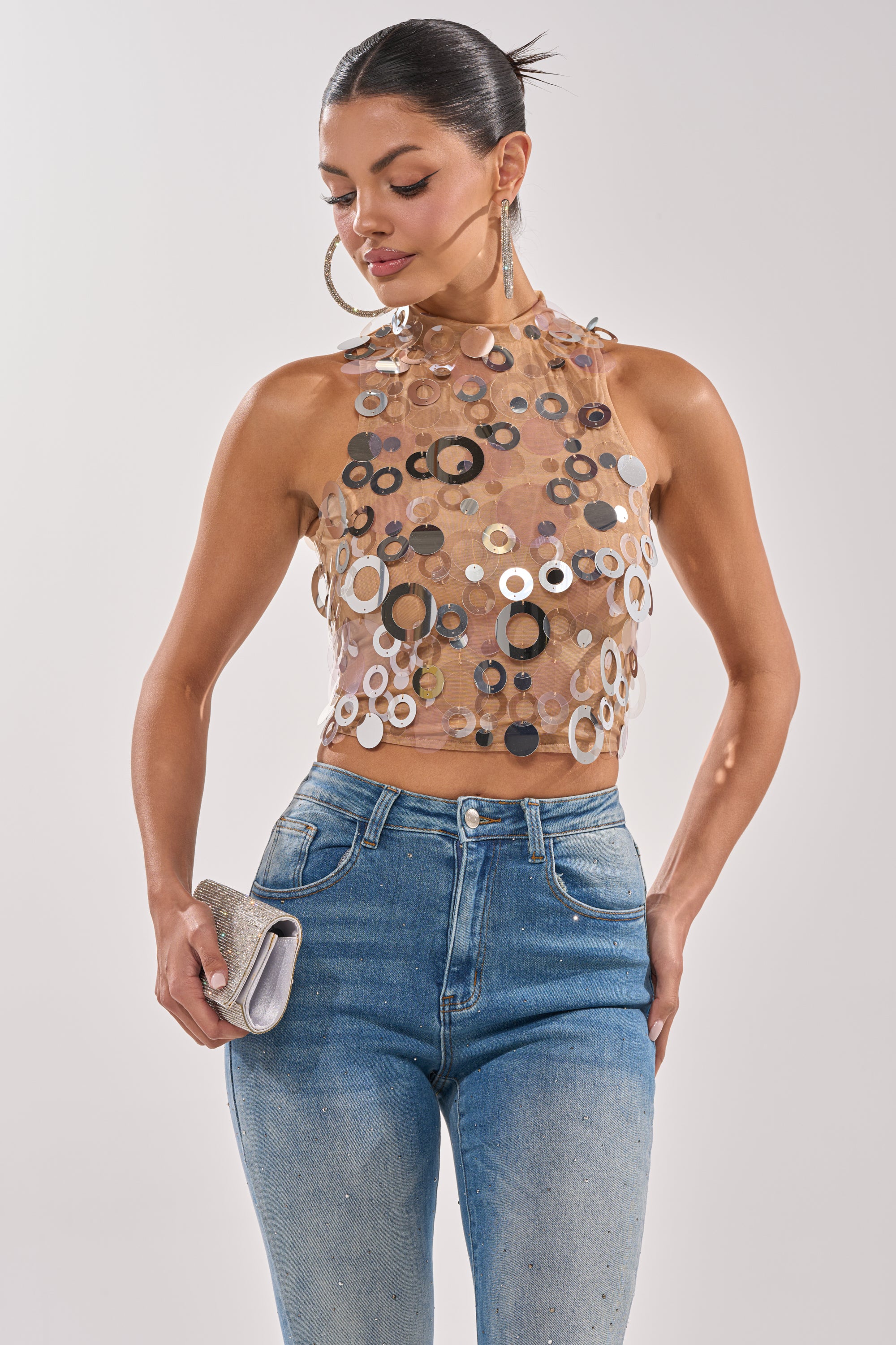 A woman stands against a plain light background wearing the BLING BLING TANK with large metallic disc embellishments, high-waisted blue jeans, hoop earrings, and holding a silver clutch. 1
