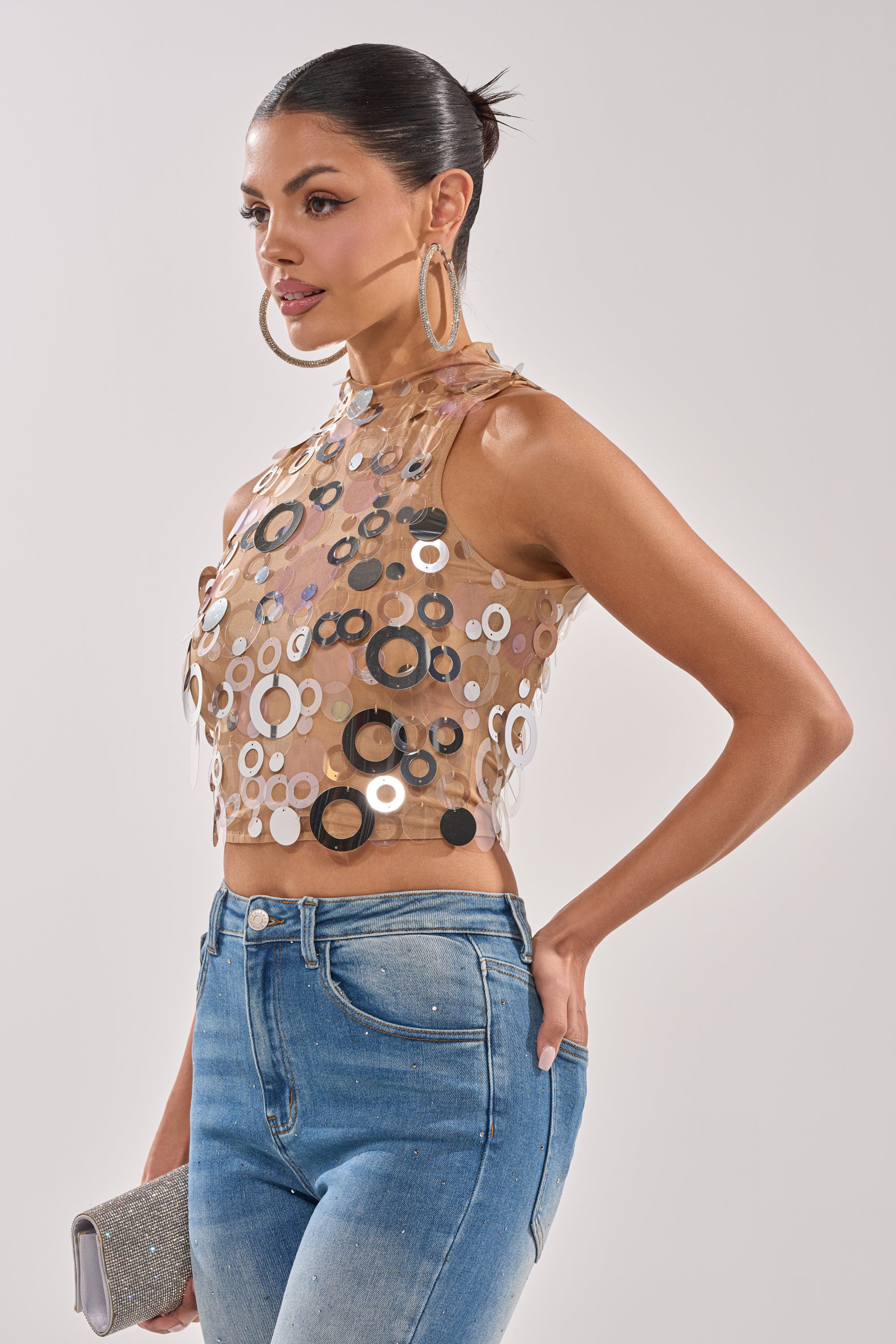 A woman with slicked-back hair wears large hoop earrings, high-waisted blue jeans, and the BLING BLING TANK—a party-ready sequin top. She holds a silver clutch and poses against a plain light background. 2