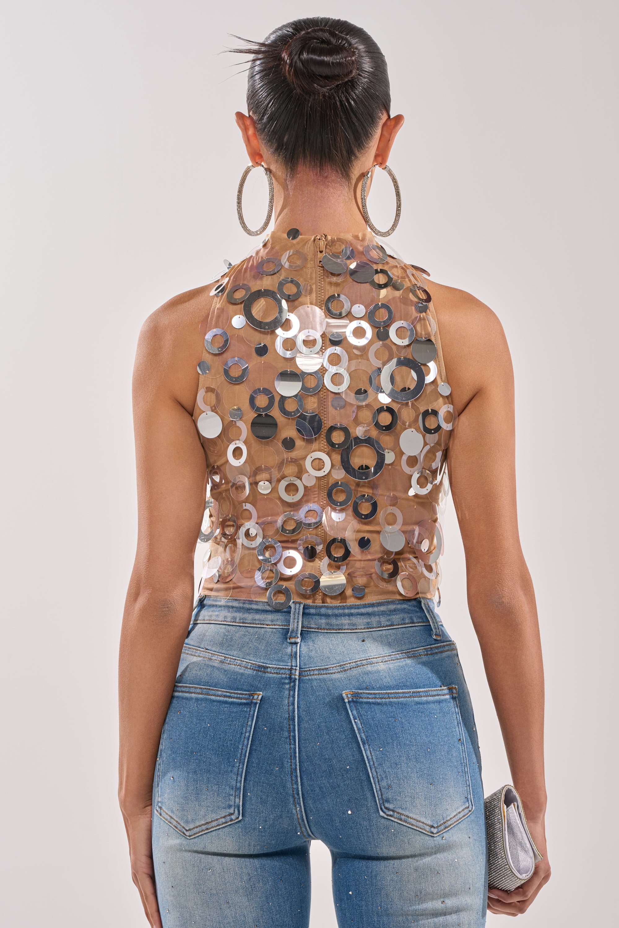 A woman with her hair in a bun and large hoop earrings, seen from the back, wears light blue high-waisted jeans, holds a small silver clutch, and rocks the sparkling BLING BLING TANK with sheer party-ready details. 3