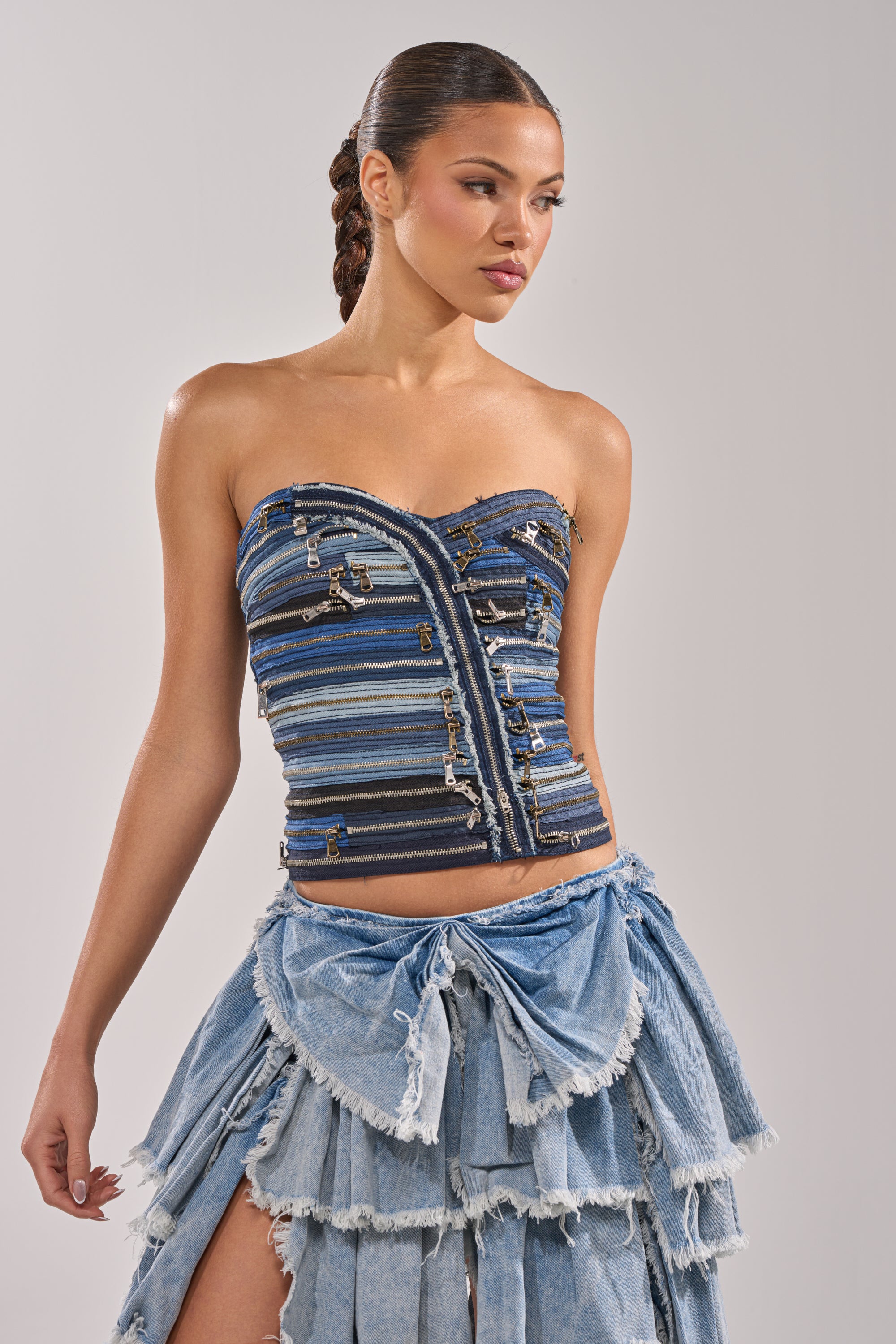 A woman models the NAME YOUR GAME ZIPPER CORSET, a strapless denim top with metallic accents and zippers, styled with a tiered ruffled light denim skirt, set against a plain light gray background for an edgy streetwear look. 0