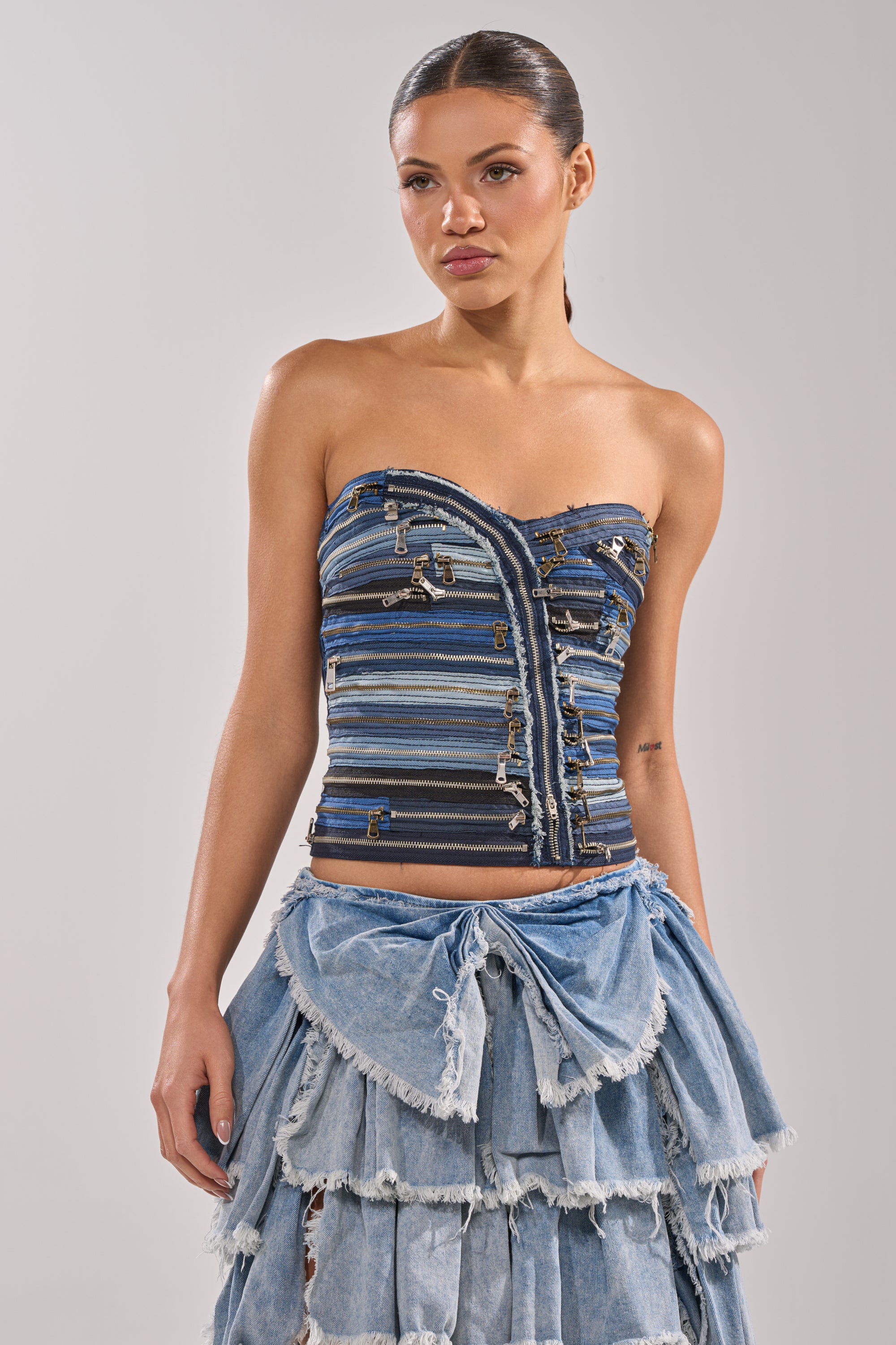 A woman stands against a light background wearing the NAME YOUR GAME ZIPPER CORSET, a strapless denim top with metal accents, paired with a layered ruffled skirt for an edgy streetwear vibe. Her hair is pulled back and she has a neutral expression. 1