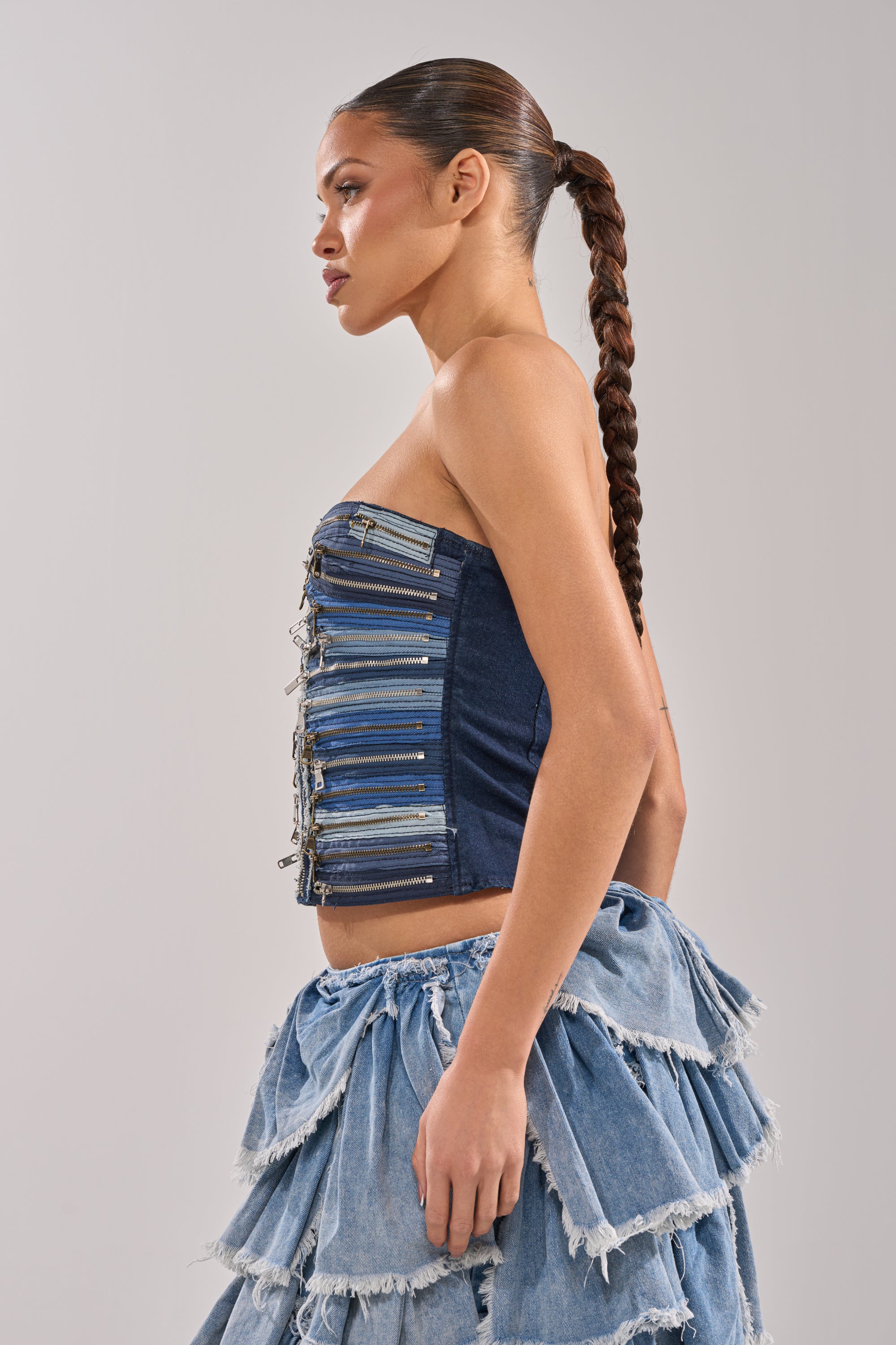 A woman with a braided ponytail poses in profile, wearing the NAME YOUR GAME ZIPPER CORSET—a strapless streetwear denim top with metal zipper details—paired with a blue denim skirt featuring layered ruffled tiers on a plain, light backdrop. 3
