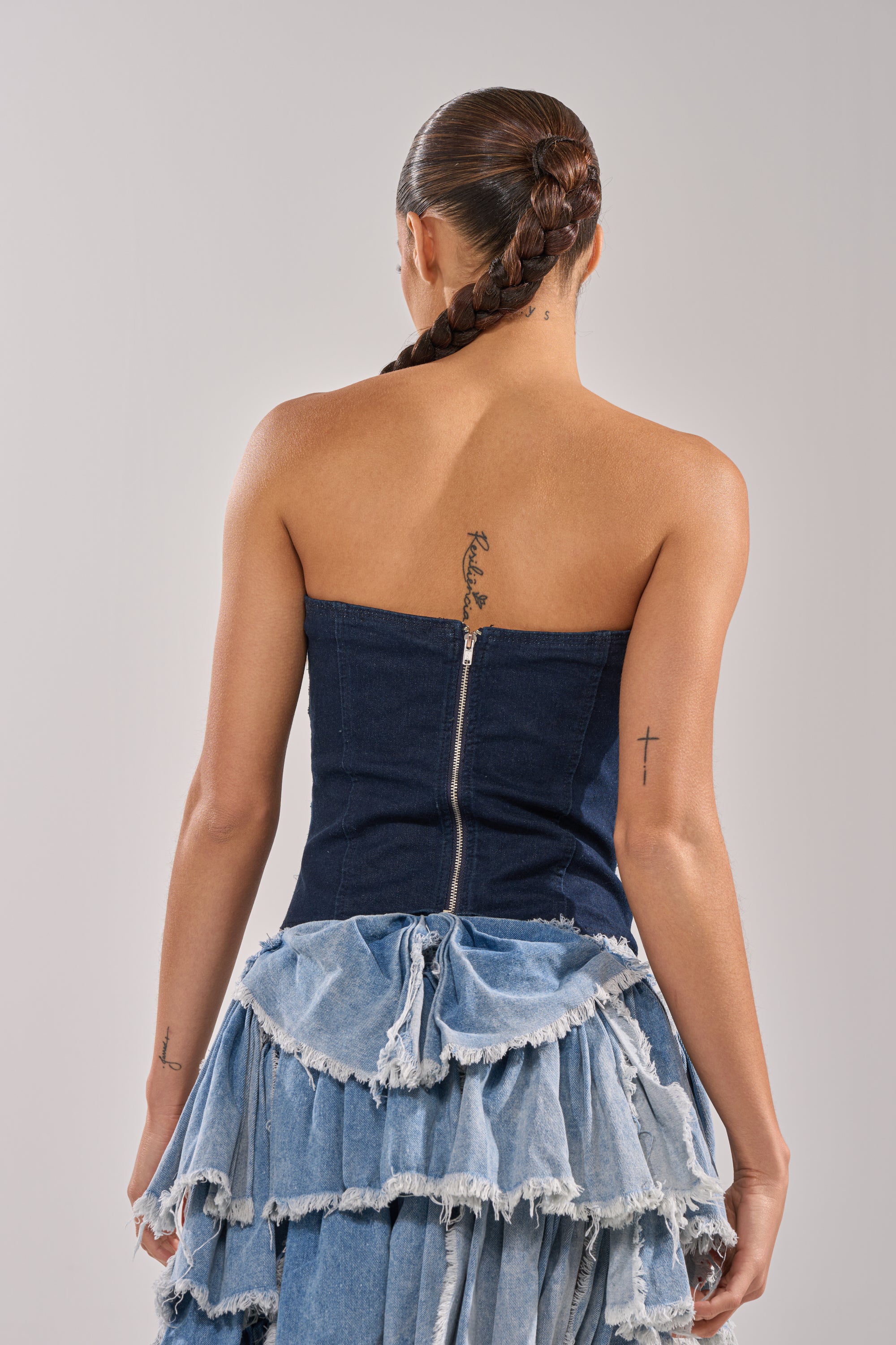 A woman with a long braided ponytail and back tattoos wears the NAME YOUR GAME ZIPPER CORSET, a strapless denim dress with ruffled skirt—perfect for edgy streetwear or making a statement at festivals against a neutral backdrop. 4