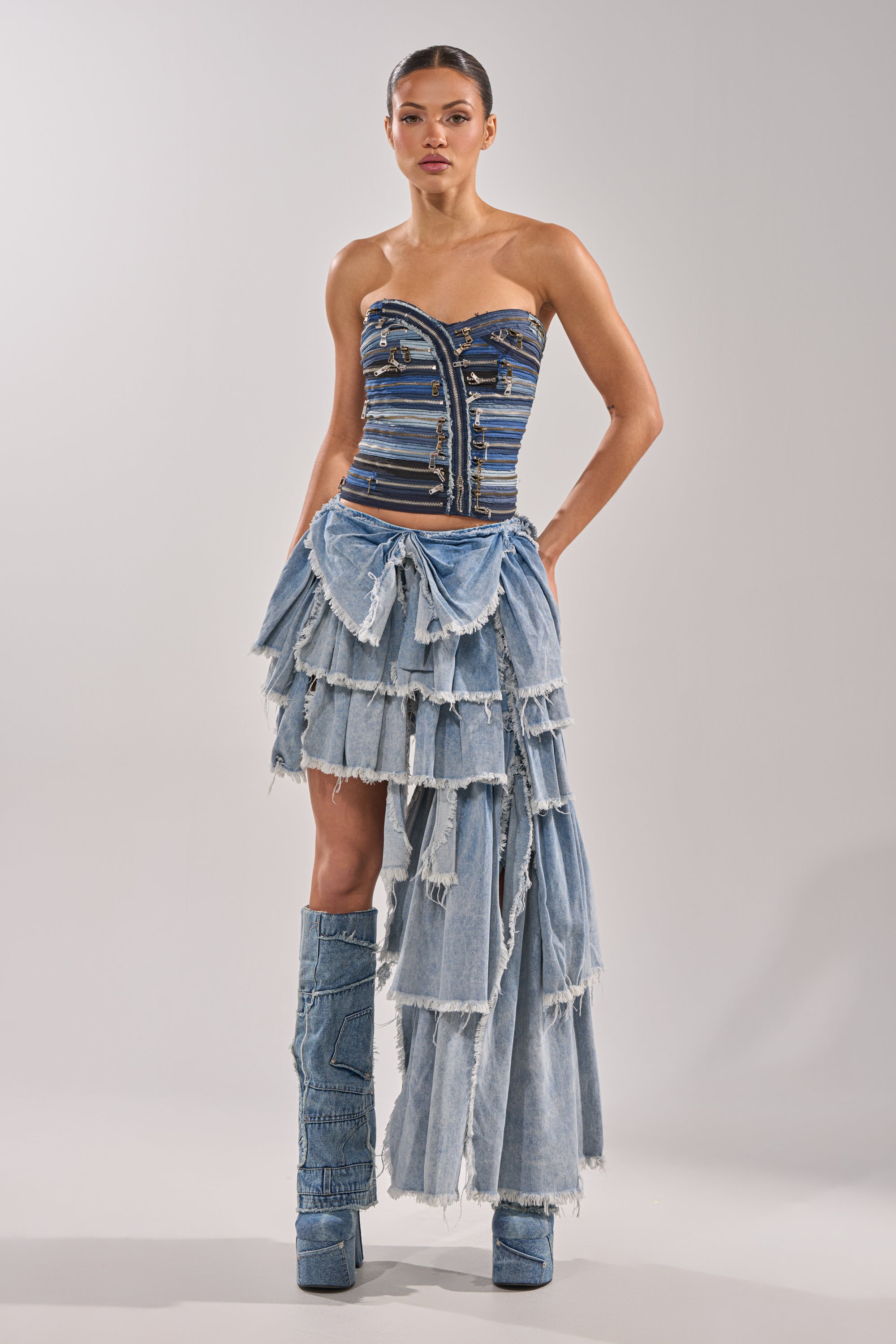 A woman wears the NAME YOUR GAME ZIPPER CORSET—a strapless denim piece with visible seams and zipper details—styled with an asymmetrical frayed denim skirt and matching knee-high boots against a plain light gray backdrop. 5