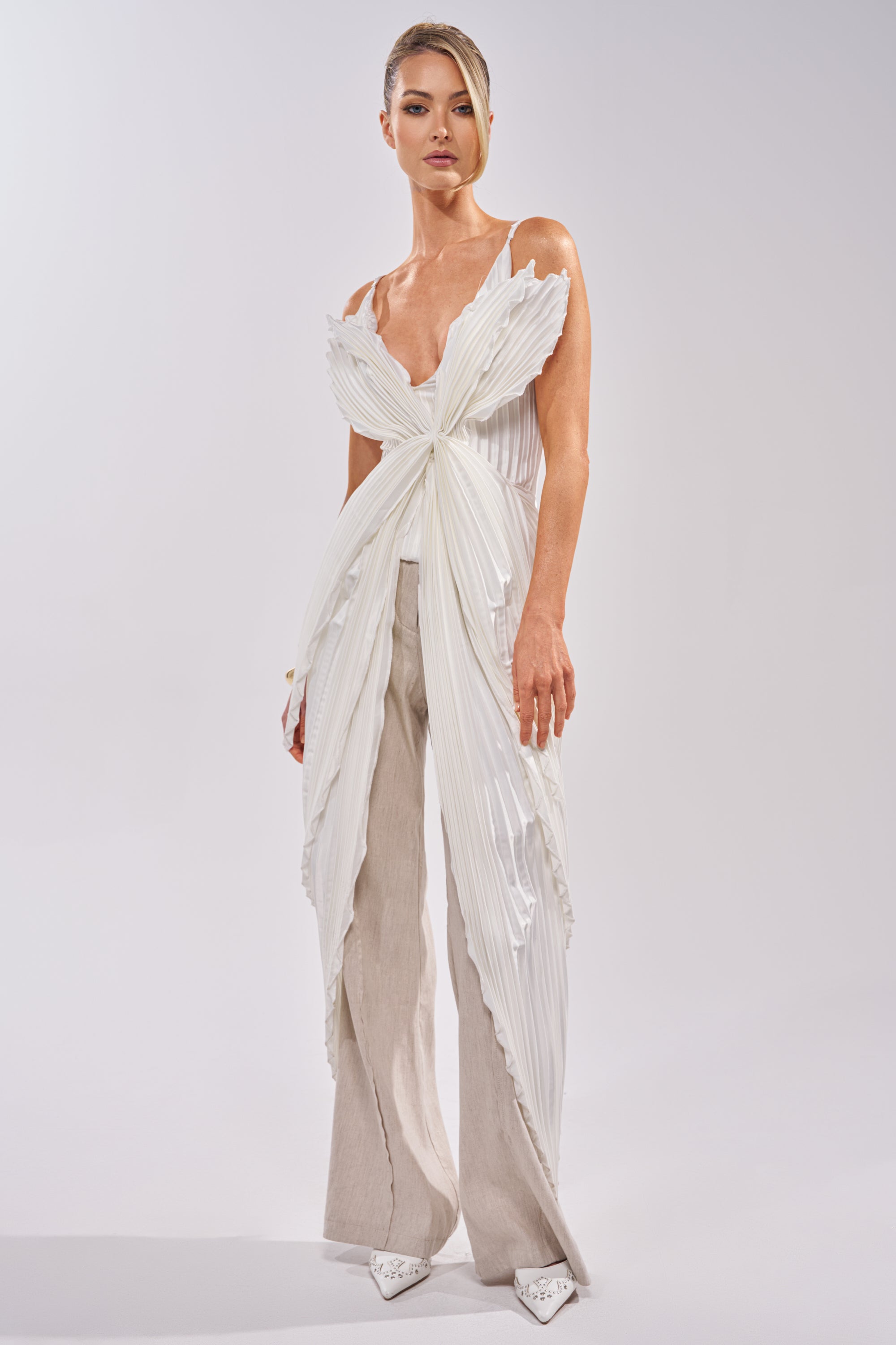 A person wears the FLYING FREE LAYERED TOP, an avant-garde satin white piece with pleated, bow-like panels, paired with light beige wide-leg pants and pointed white shoes against a plain background. 0
