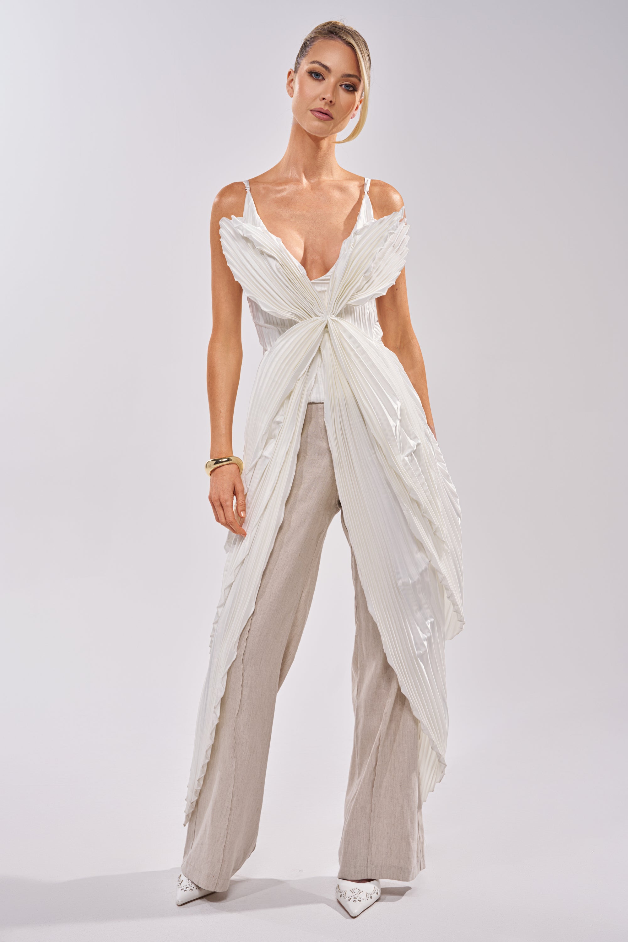 A woman stands confidently in the FLYING FREE LAYERED TOP—satin white with pleated panels and a large front bow—paired with loose beige pants and white pointed-toe heels, set against a plain light background. 1