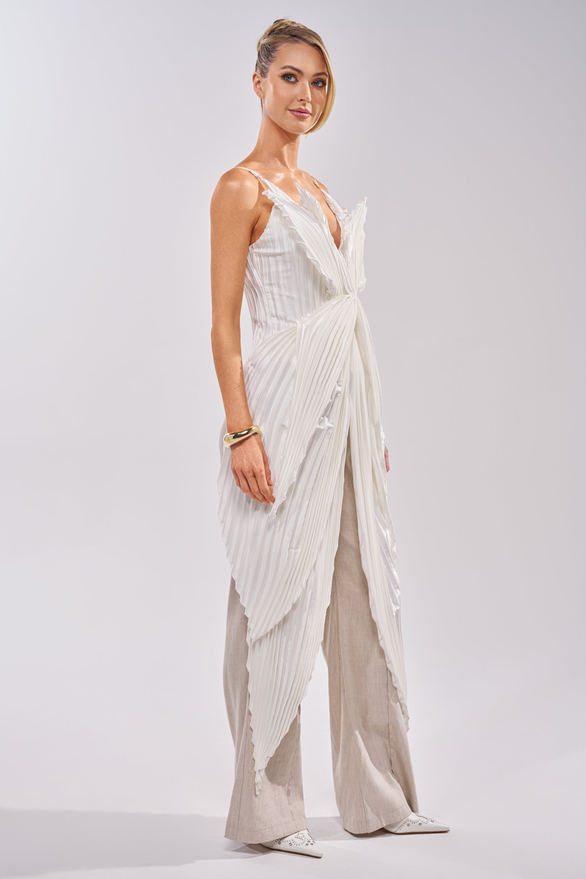 A woman stands confidently in the FLYING FREE LAYERED TOP, styled over beige wide-leg pants with a gold bracelet and white pointed shoes, posed against a simple light gray background. 2