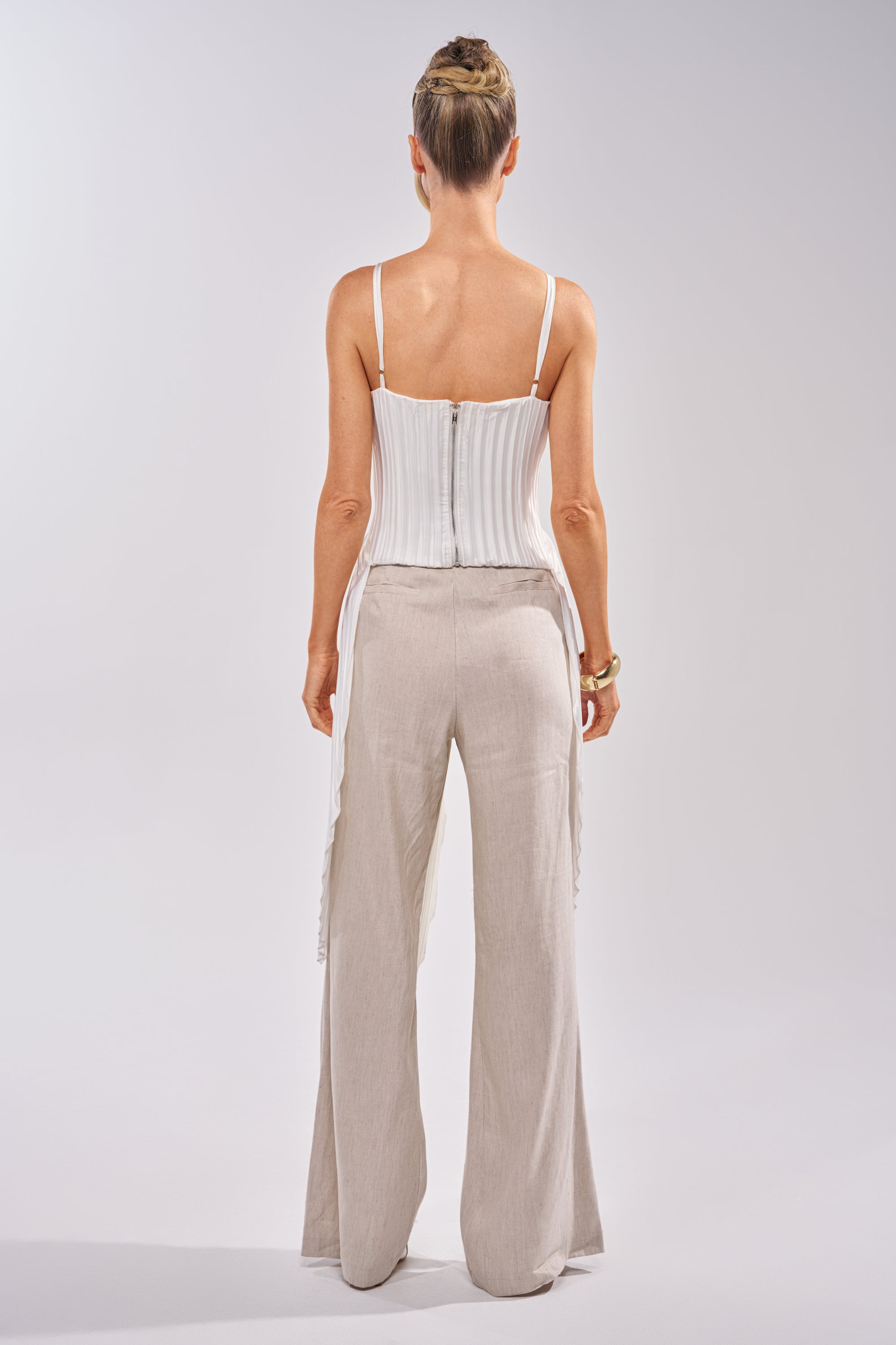 A person faces away from the camera wearing the FLYING FREE LAYERED TOP in satin white with pleated panels, paired with loose light beige pants. Their hair is in a neat bun against a plain, light background. 3