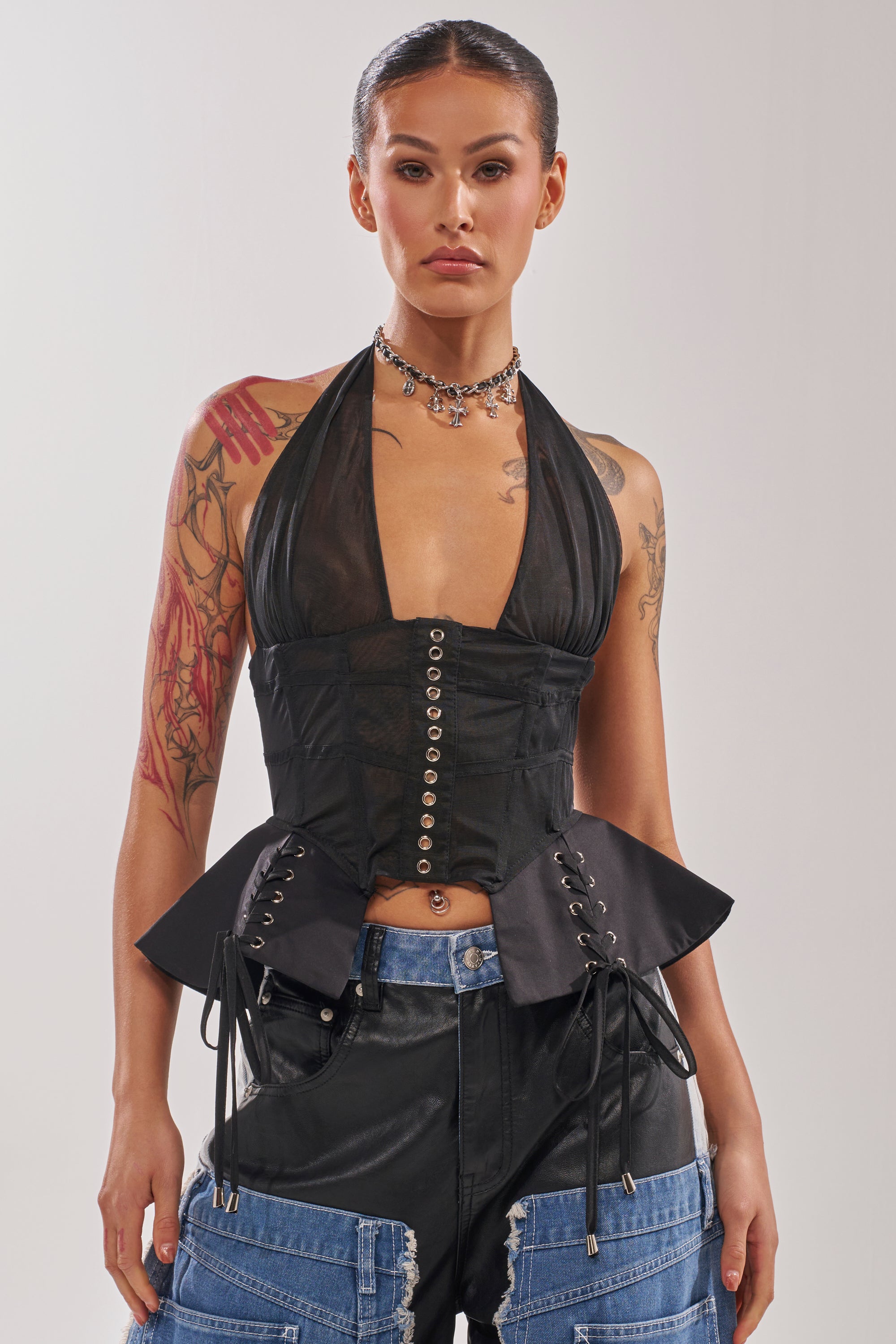 A woman with dark hair tied back wears the DREAM GIRL SHEER CORSET, featuring boned construction and lace-up details, paired with black pants with denim pockets. She accessorizes with a silver choker and has visible arm tattoos. 0