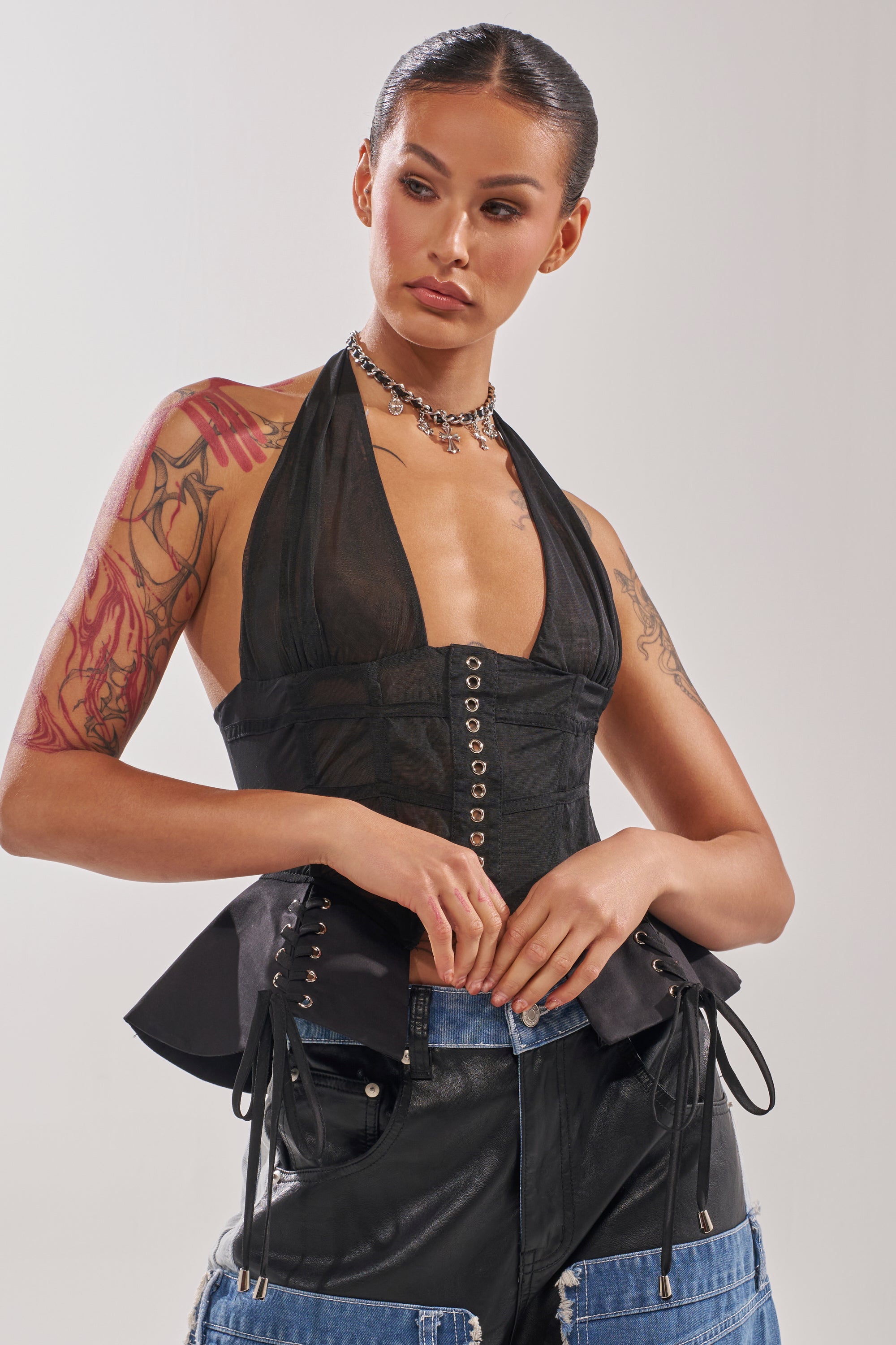 A woman with tattoos wears the DREAM GIRL SHEER CORSET, styled with layered necklaces and two-toned denim-leather pants, standing against a light background and gazing to the side with relaxed arms. 1