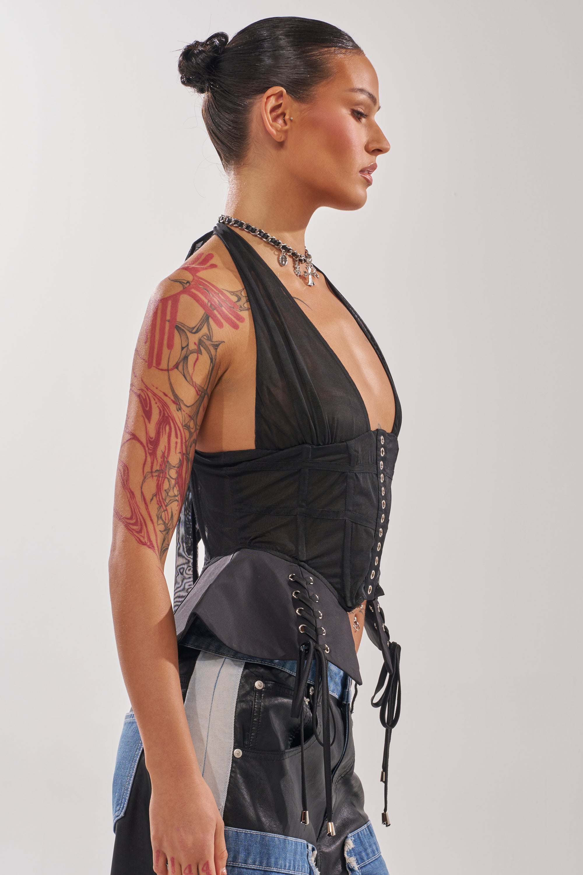 A woman with slicked-back hair and visible arm tattoos models the DREAM GIRL SHEER CORSET, featuring boned construction and a lace-up front, paired with layered necklaces and two-tone jeans, in profile against a plain backdrop. 3