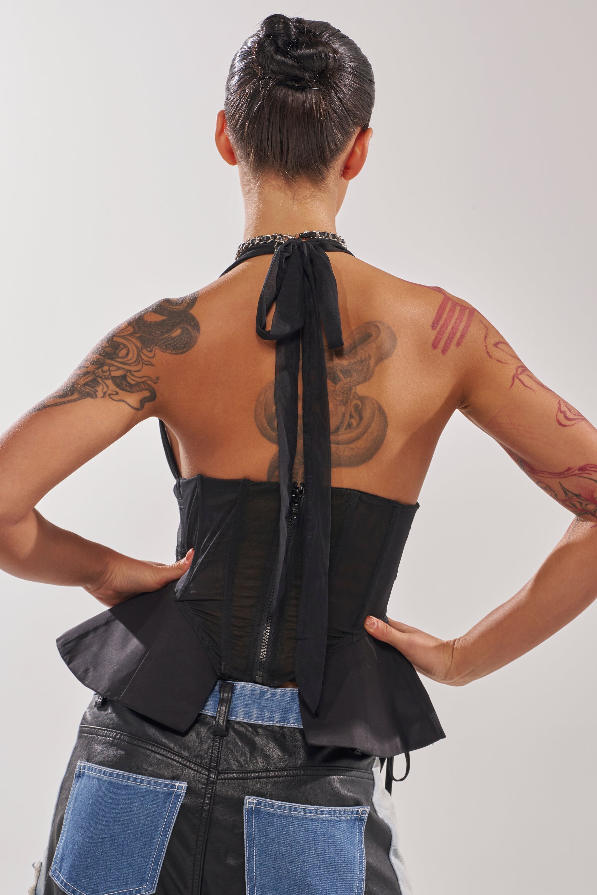 A person with short dark hair and arm and back tattoos stands hands on hips, facing away from the camera against a plain background, wearing the DREAM GIRL SHEER CORSET in black and denim patchwork pants. 4