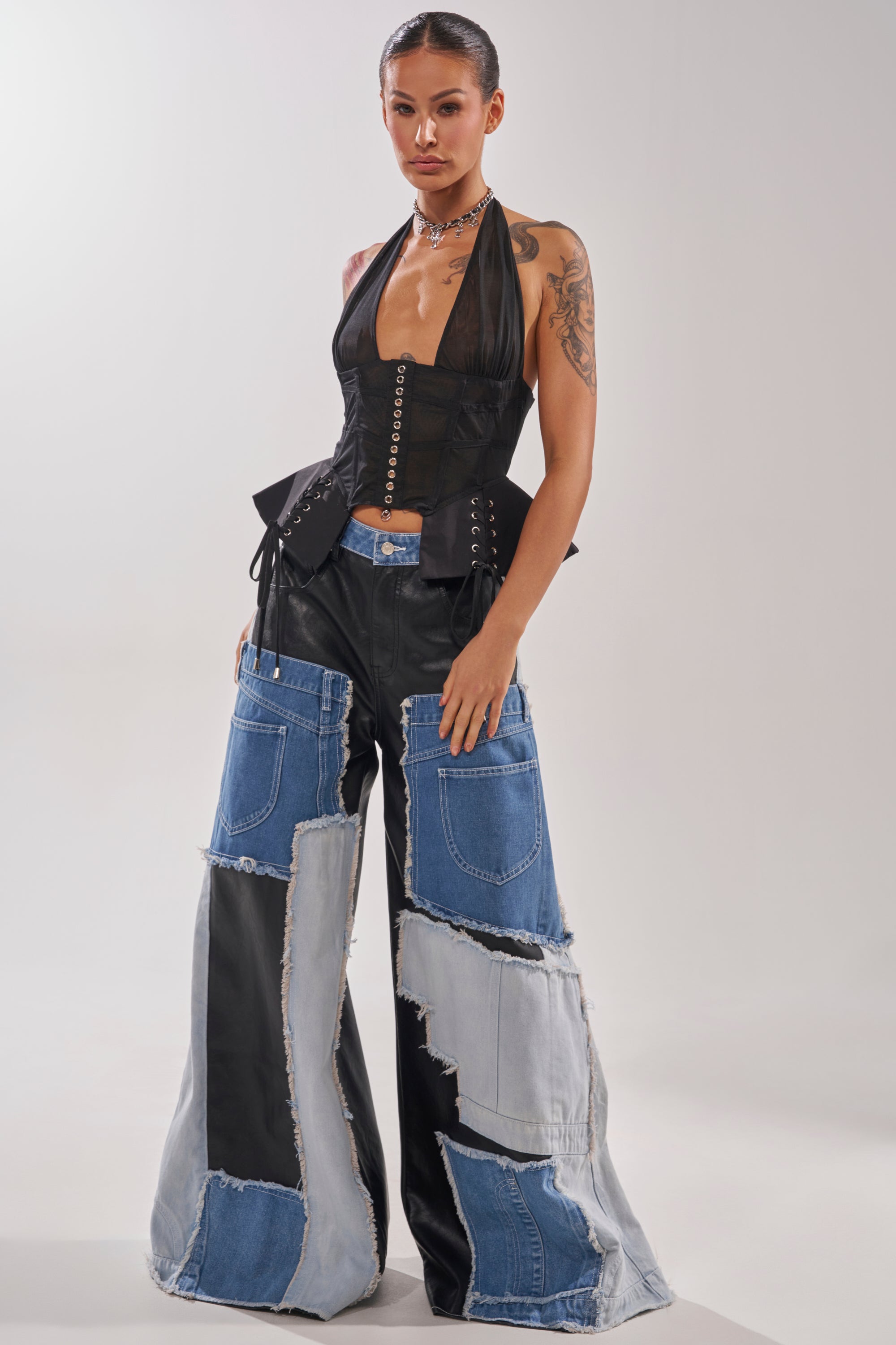 A woman poses in the DREAM GIRL SHEER CORSET, a black boned corset top, paired with wide-leg patchwork denim jeans with large pockets and frayed edges. She has slicked-back hair, a tattoo, and layered necklaces. 6