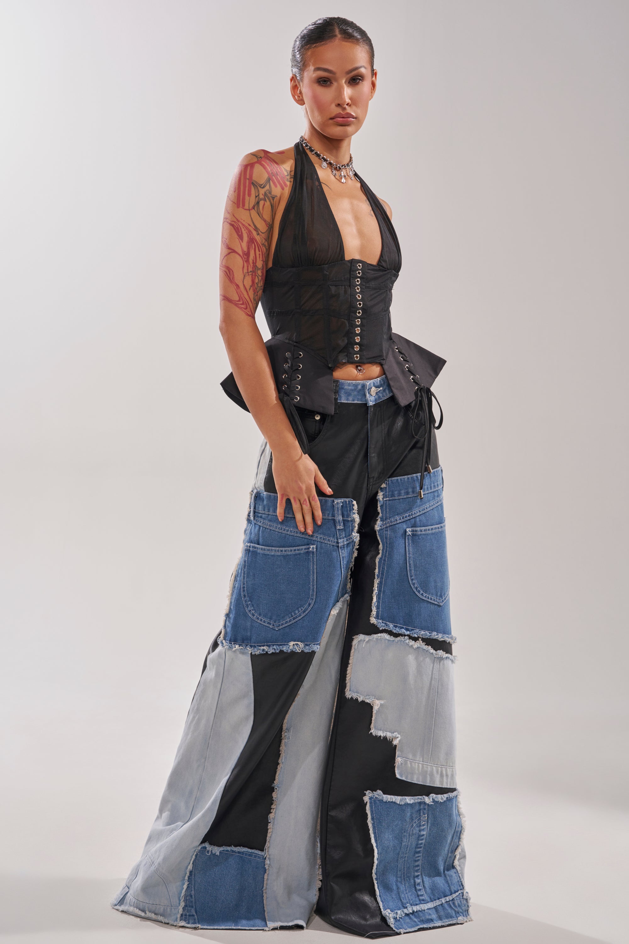 A woman stands confidently in the DREAM GIRL SHEER CORSET with boned construction, paired with wide-leg patchwork denim pants, layered necklaces, and slicked-back hair. She has an arm tattoo and poses against a plain light background. 8