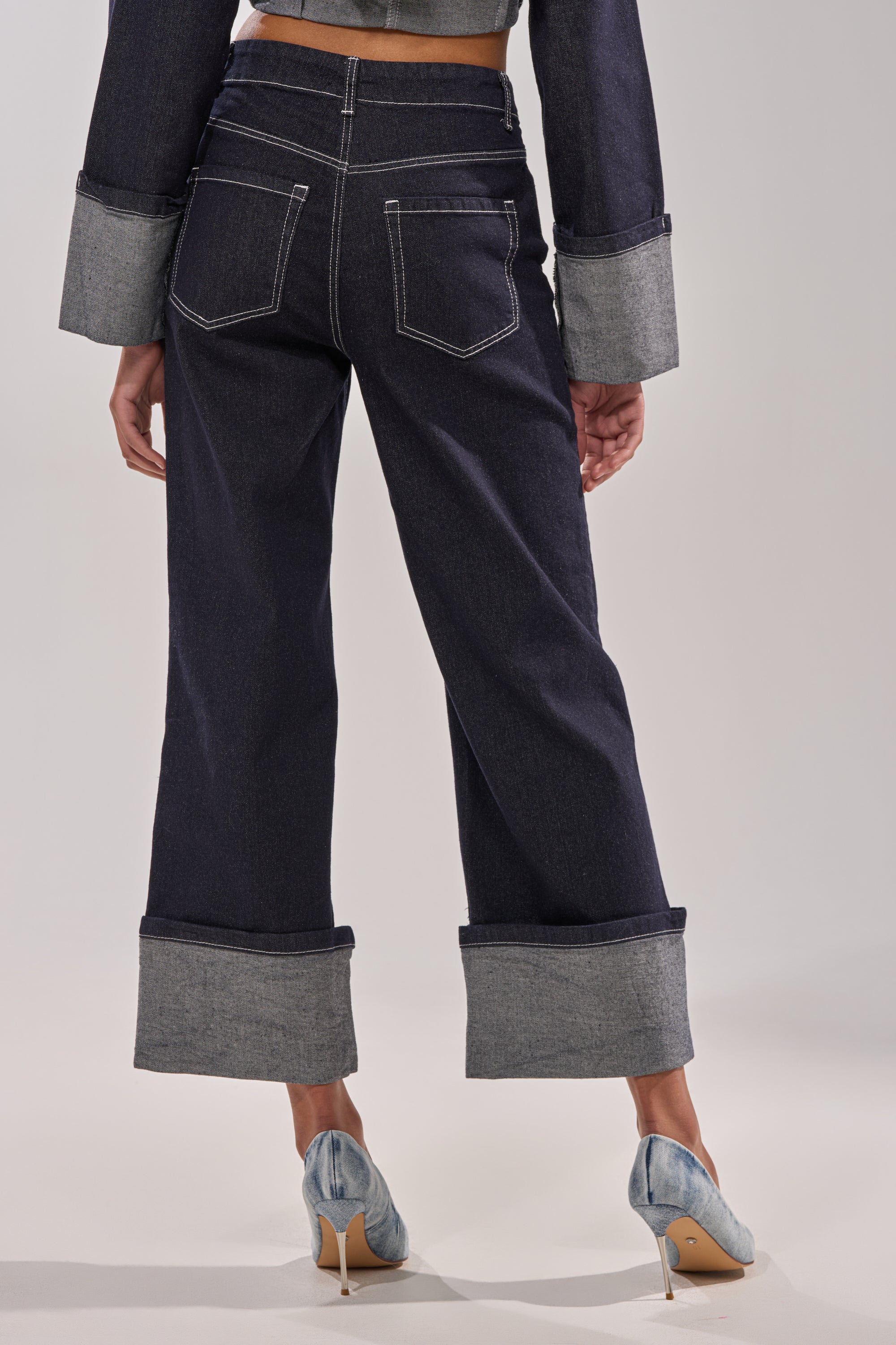 A person faces away in DON'T TALK TO ME WIDE LEG DENIM JEANS, featuring dark wash, wide legs, large cuffed hems, and white stitching. They're paired with light blue heeled shoes against a plain background. 5