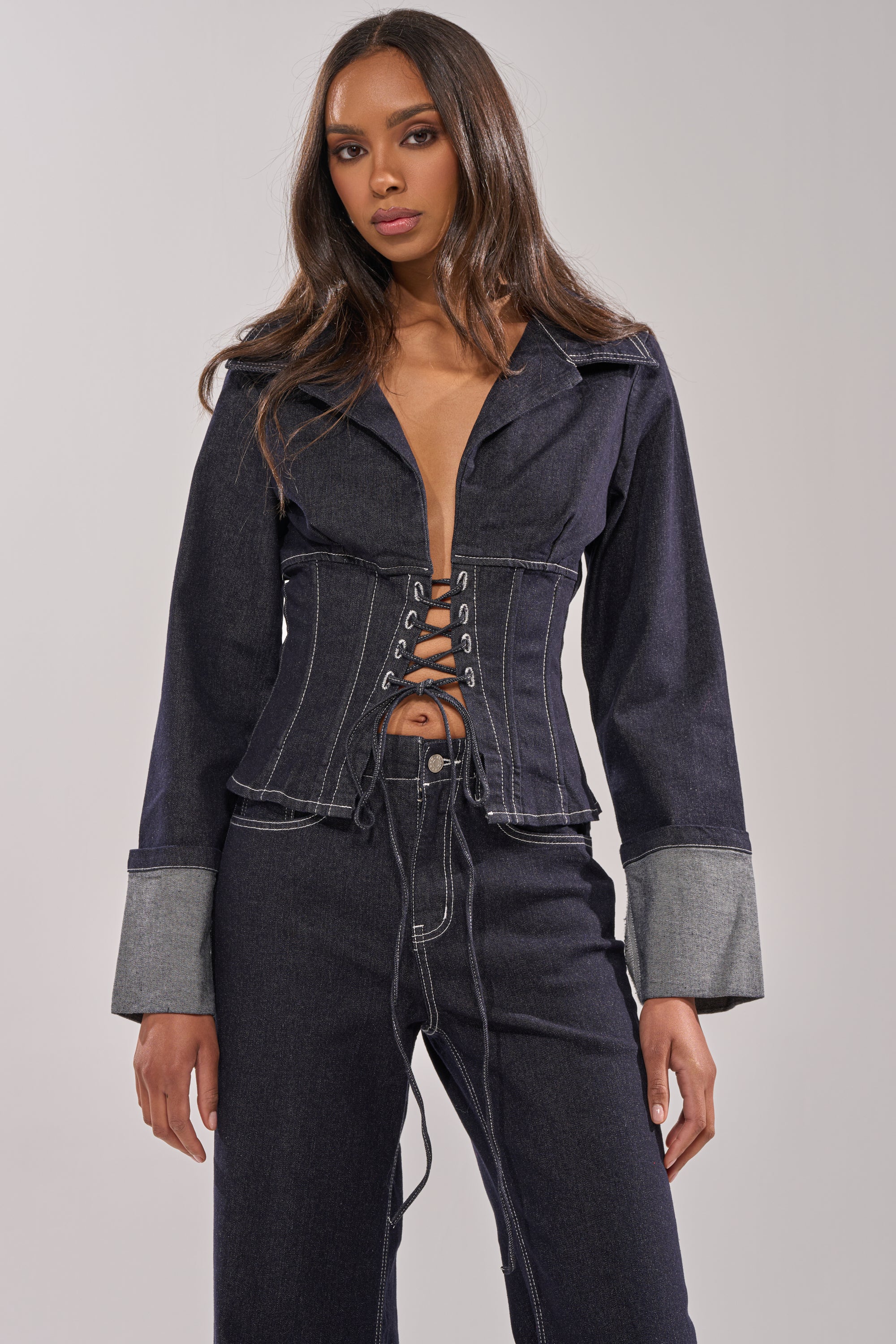A woman in the DON'T TALK TO ME LACE UP DENIM BLOUSE, featuring tailored wide cuffed sleeves and a corset-style lace-up front, stands against a plain background with a serious expression. 0
