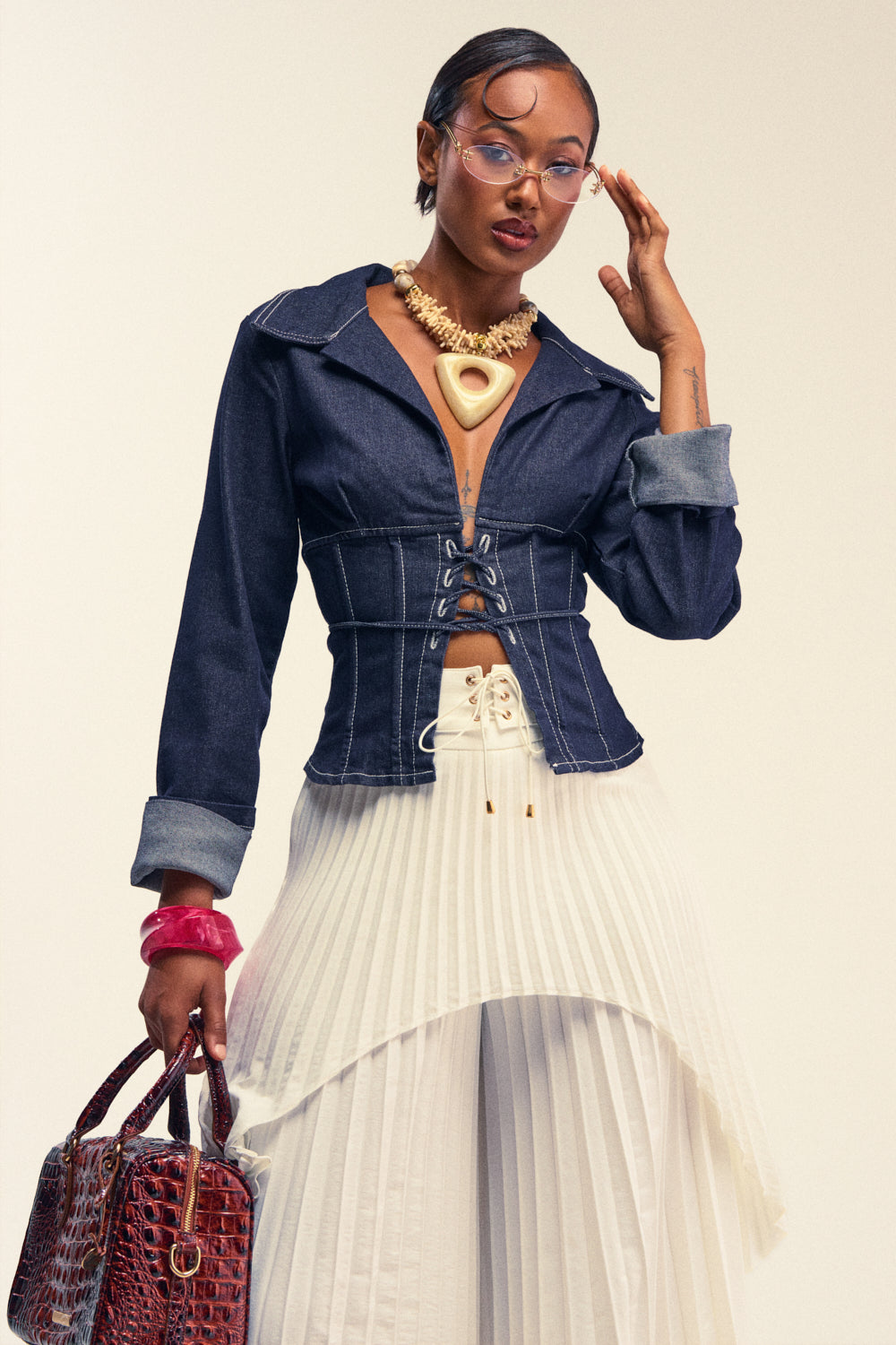 A woman poses in the DON'T TALK TO ME LACE UP DENIM BLOUSE—a tailored, dark-wash denim jacket featuring a corset-inspired lace-up detail—paired with a white pleated skirt, large gold necklace, and clear glasses. She holds a textured brown handbag in one hand while touching her face with the other.