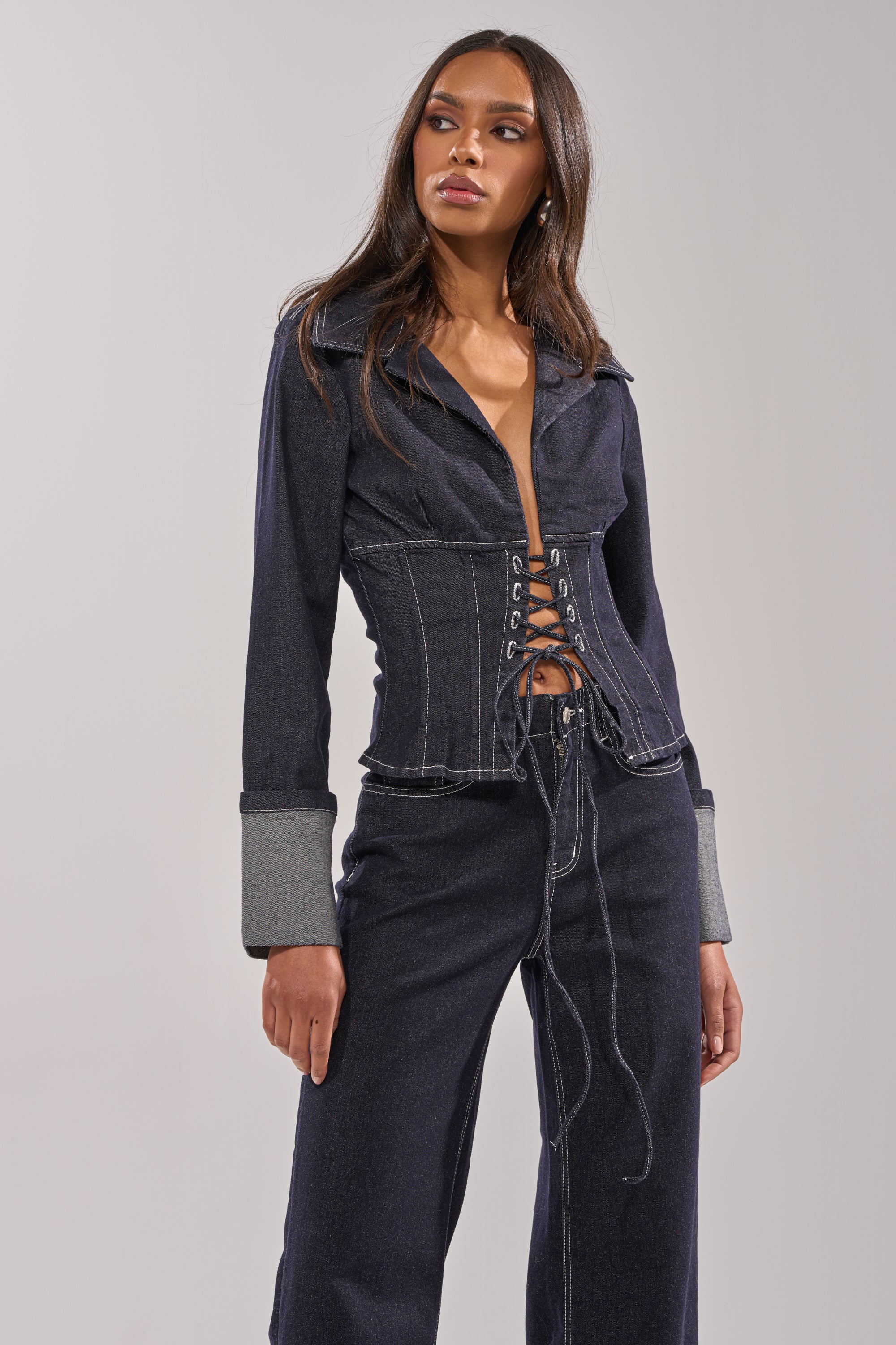 A woman wears the DON'T TALK TO ME LACE UP DENIM BLOUSE featuring a corset-inspired lace-up front and wide cuffs, paired with matching denim pants, standing against a plain backdrop and looking confidently to the side. 1