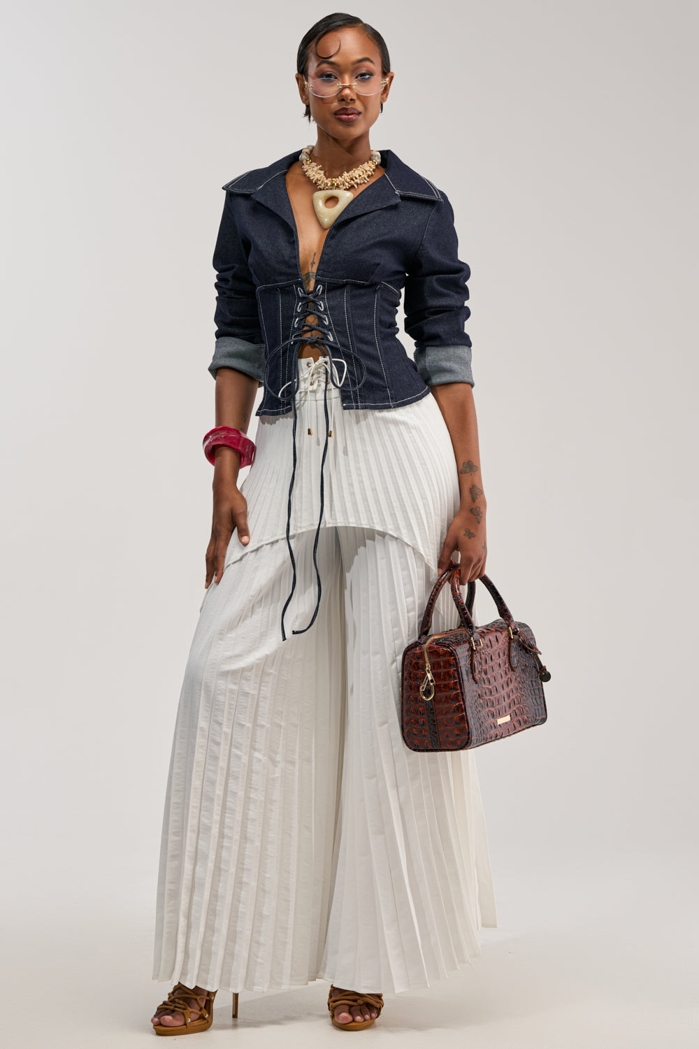 A stylish woman wears the DON'T TALK TO ME LACE UP DENIM BLOUSE, tailored in a dark wash, paired with pleated white wide-leg pants, heels, and bold jewelry. She holds a brown textured handbag and stands confidently against a neutral background.