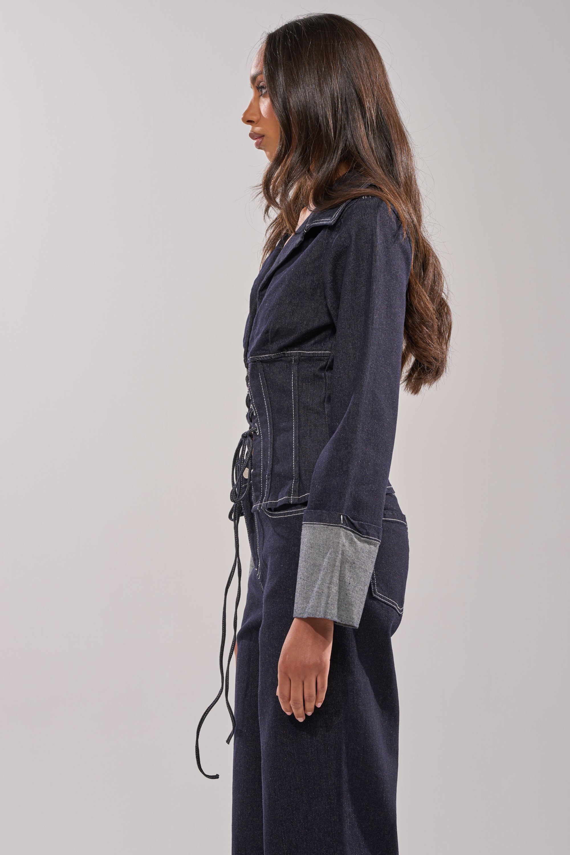 A woman with long brown hair stands in profile, wearing the DON'T TALK TO ME LACE UP DENIM BLOUSE with a corset-inspired lace-up front and wide cuffed sleeves, paired with matching denim pants against a plain light background. 2