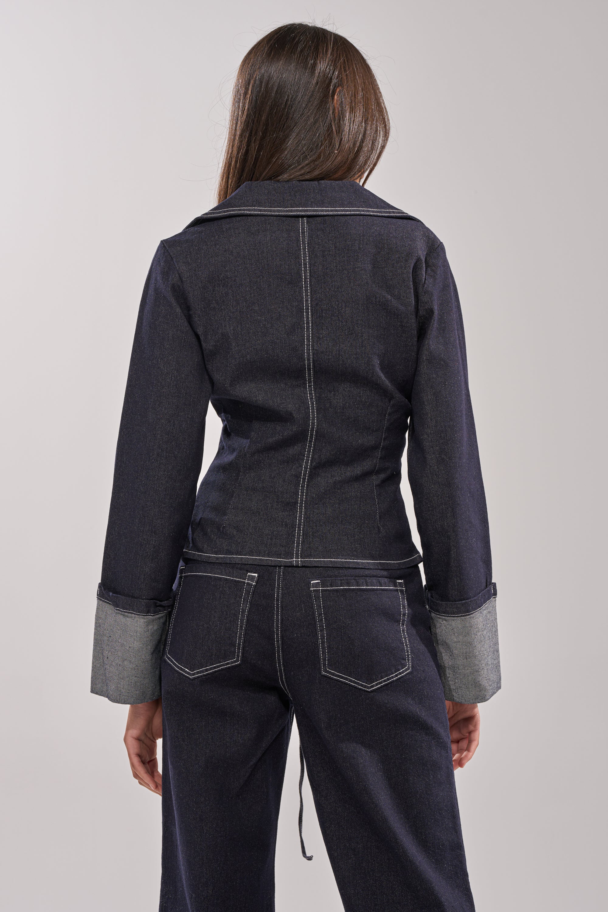 A person with long brown hair is shown from the back wearing the DON'T TALK TO ME LACE UP DENIM BLOUSE in a dark wash, featuring rolled-up sleeves and a tailored silhouette for a sleek, modern look against a plain light background. 3