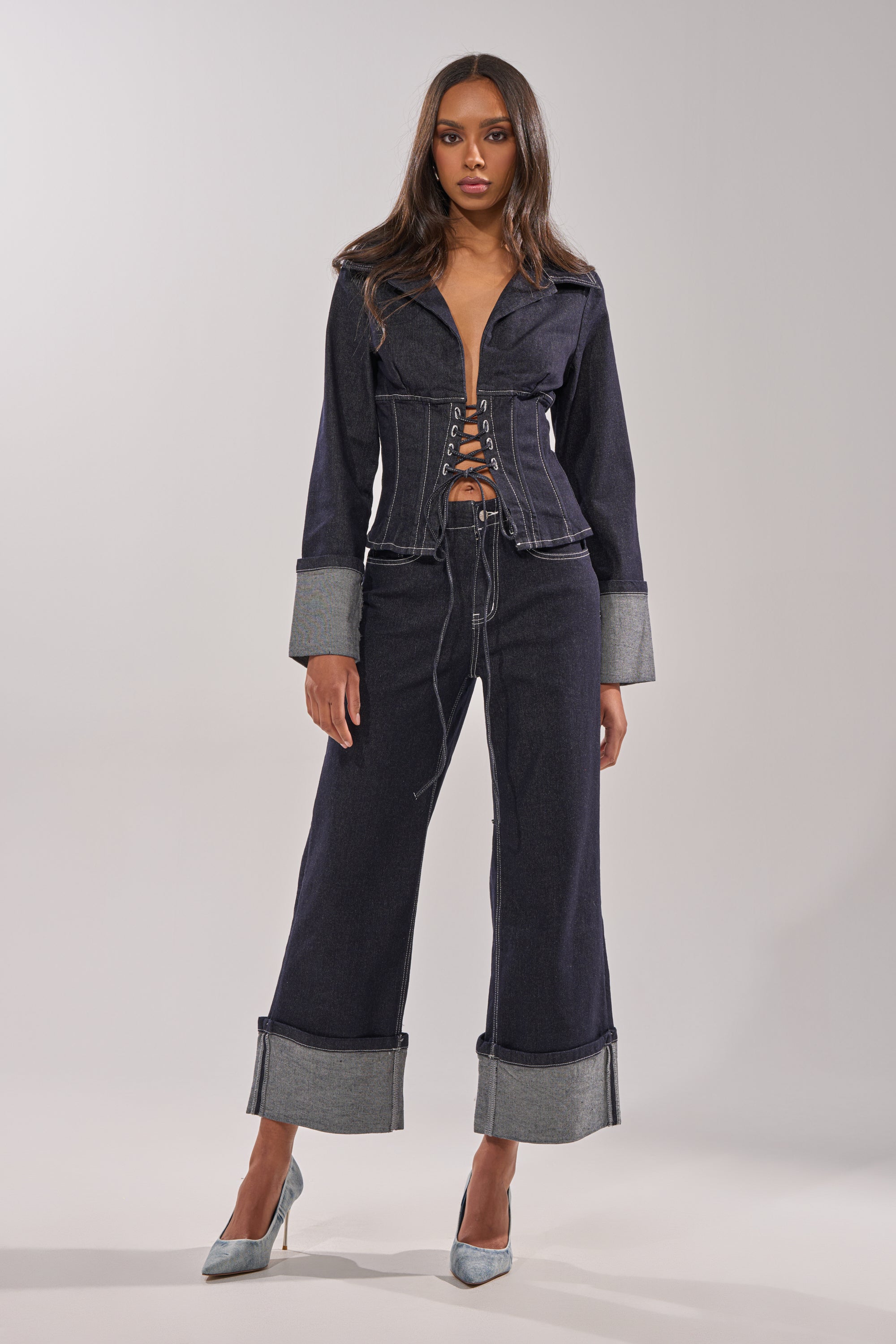 A woman poses against a plain light gray background, wearing the DON'T TALK TO ME LACE UP DENIM BLOUSE—a dark-wash, corset-inspired top—styled with wide-legged cuffed pants and gray high heels. 4