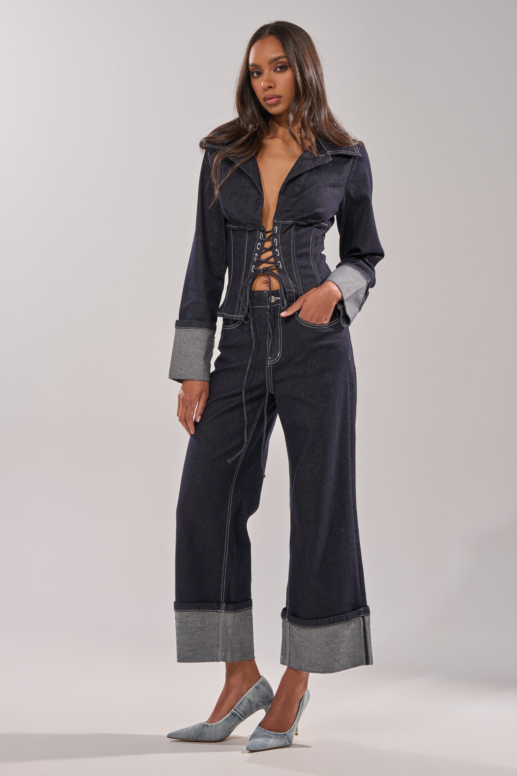 A woman poses in the DON'T TALK TO ME LACE UP DENIM BLOUSE and wide-leg, cuffed jeans. She pairs the dark-wash outfit with pointed-toe heels, wears her hair long and straight, and stands against a plain light background. 5