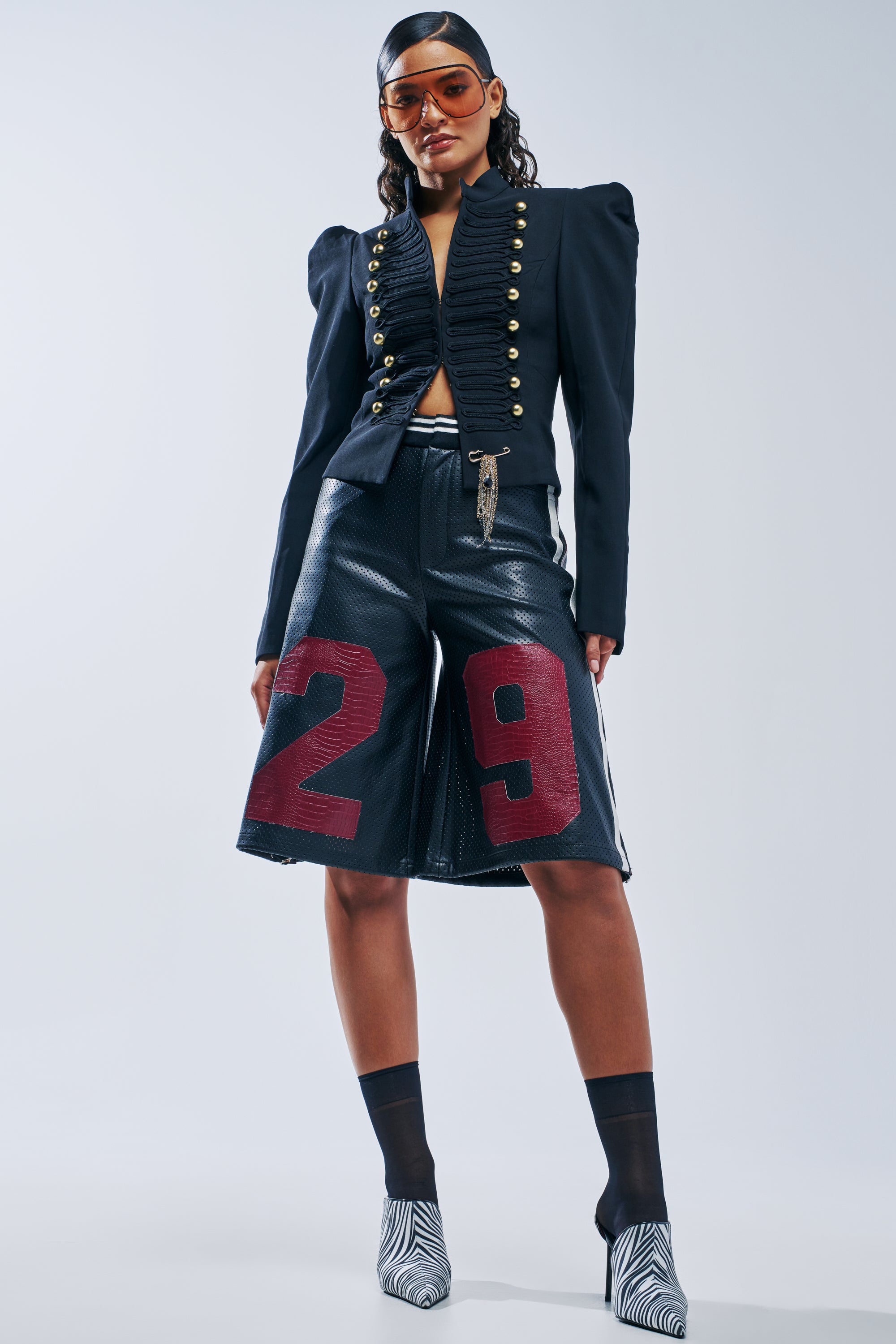 A model wearing a black military-style jacket and LUCKY 29 BERMUDA SHORT IN BLACK MULTI, paired with black socks and zebra-print heels.