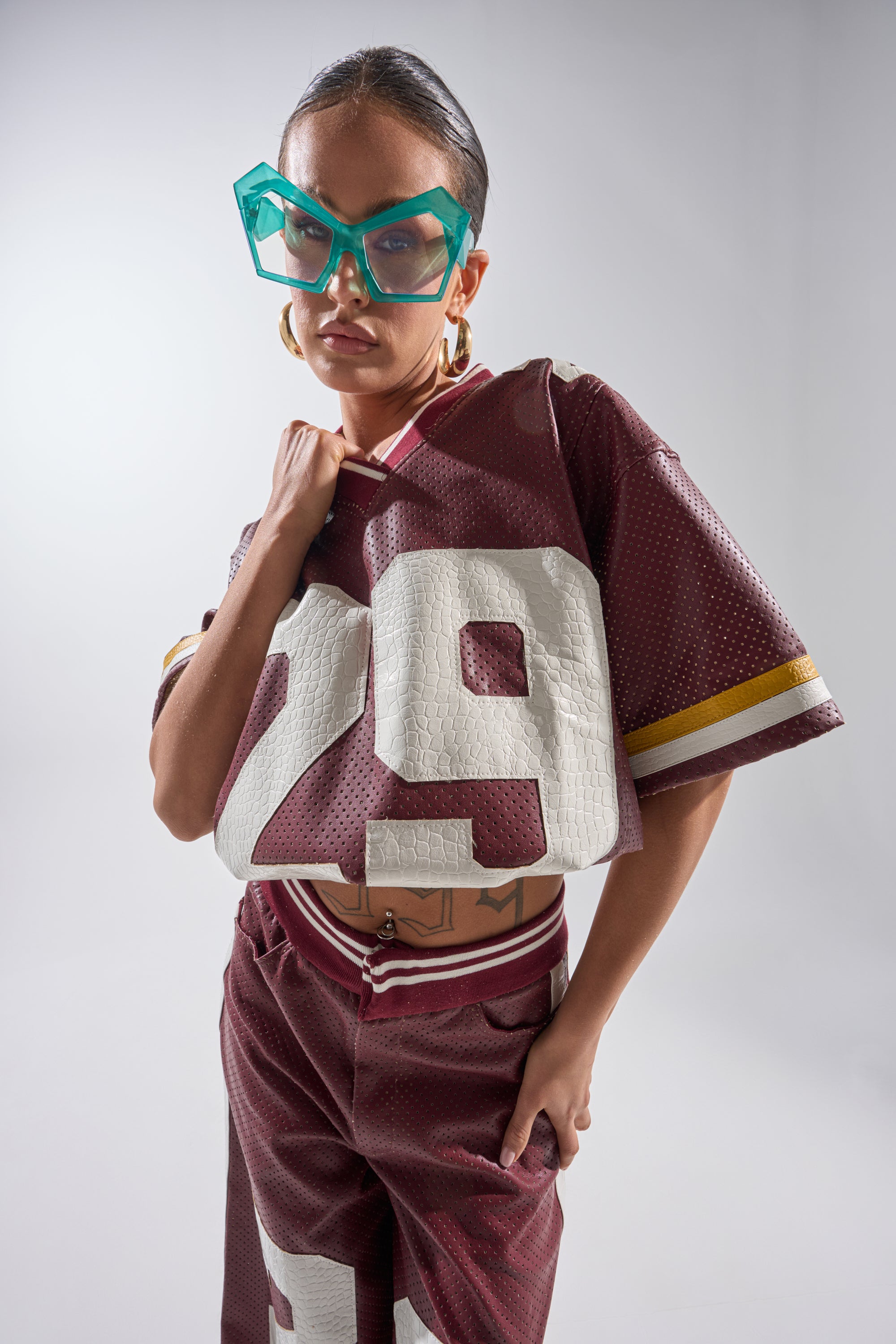 A model confidently poses against a plain light background, wearing the LUCKY 29 JERSEY in maroon faux leather with matching pants, oversized blue glasses, and large hoop earrings. 0