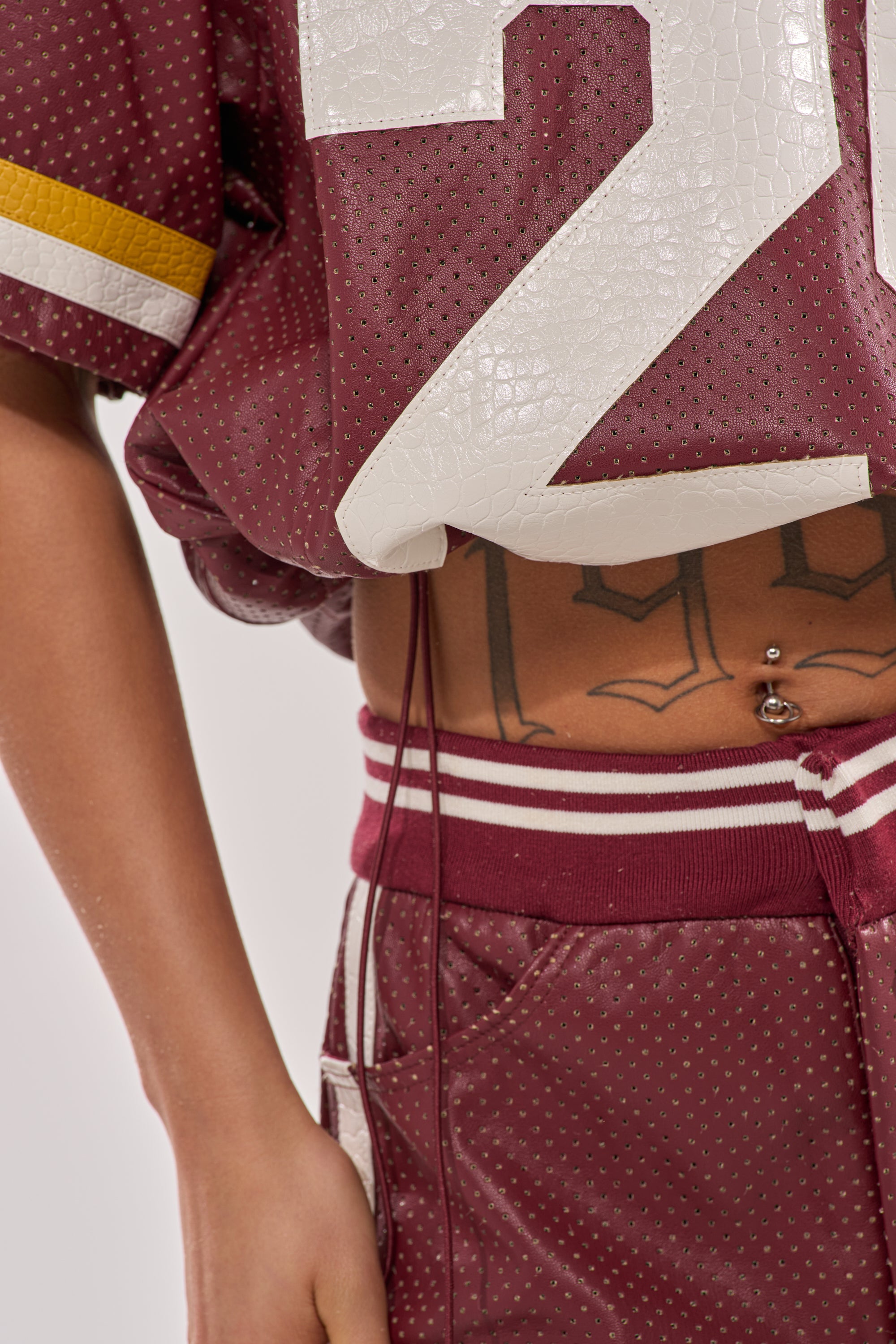 A person wearing the LUCKY 29 JERSEY and matching shorts, both in perforated burgundy with varsity-style numbers, reveals a belly tattoo and navel piercing. The image is cropped to show only the torso and part of one arm. 11