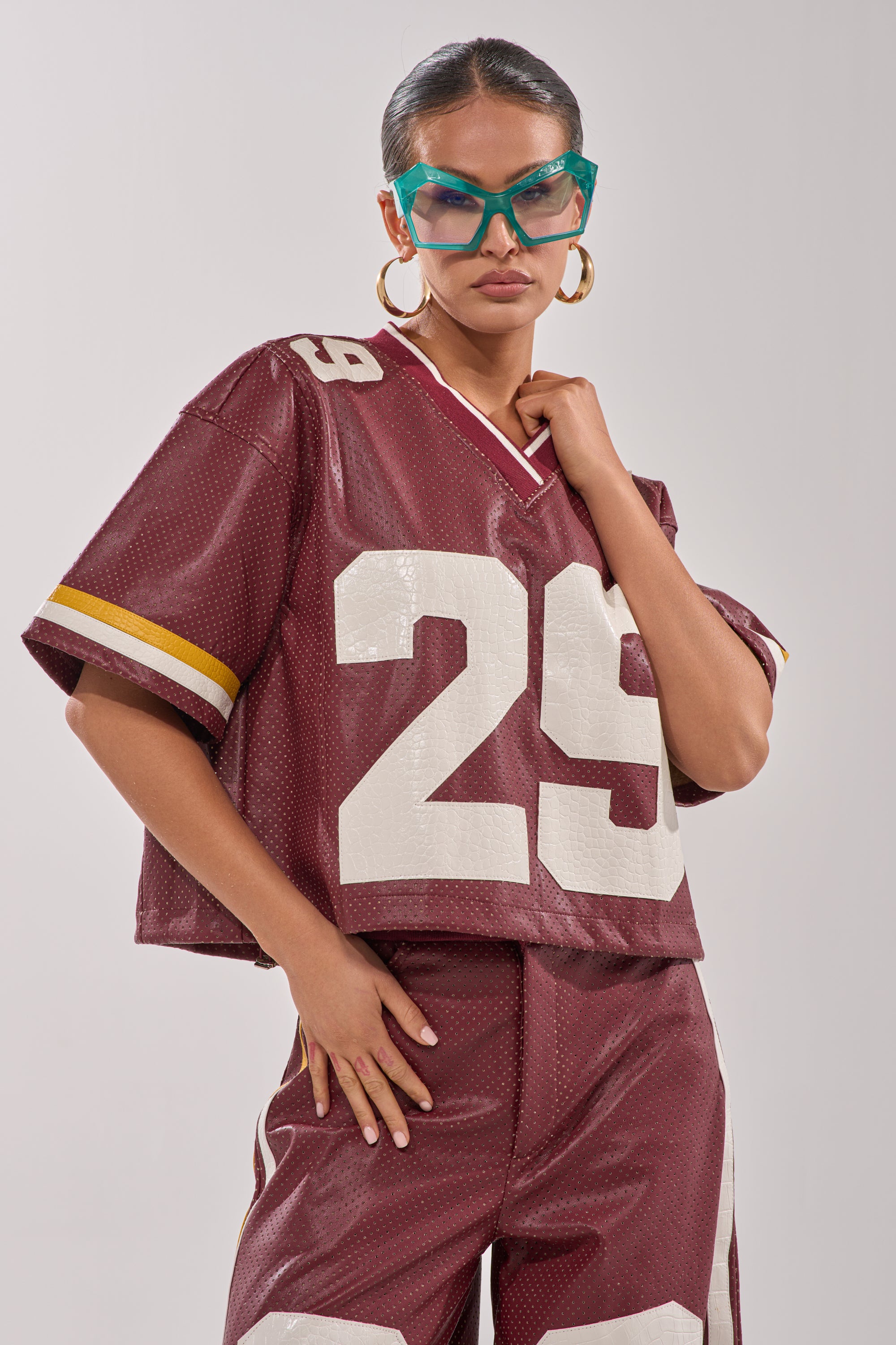 Model wears the LUCKY 29 JERSEY, a maroon faux leather piece with varsity-style number 29, paired with matching pants, large hoop earrings, and teal sunglasses. She poses confidently against a plain background. 3