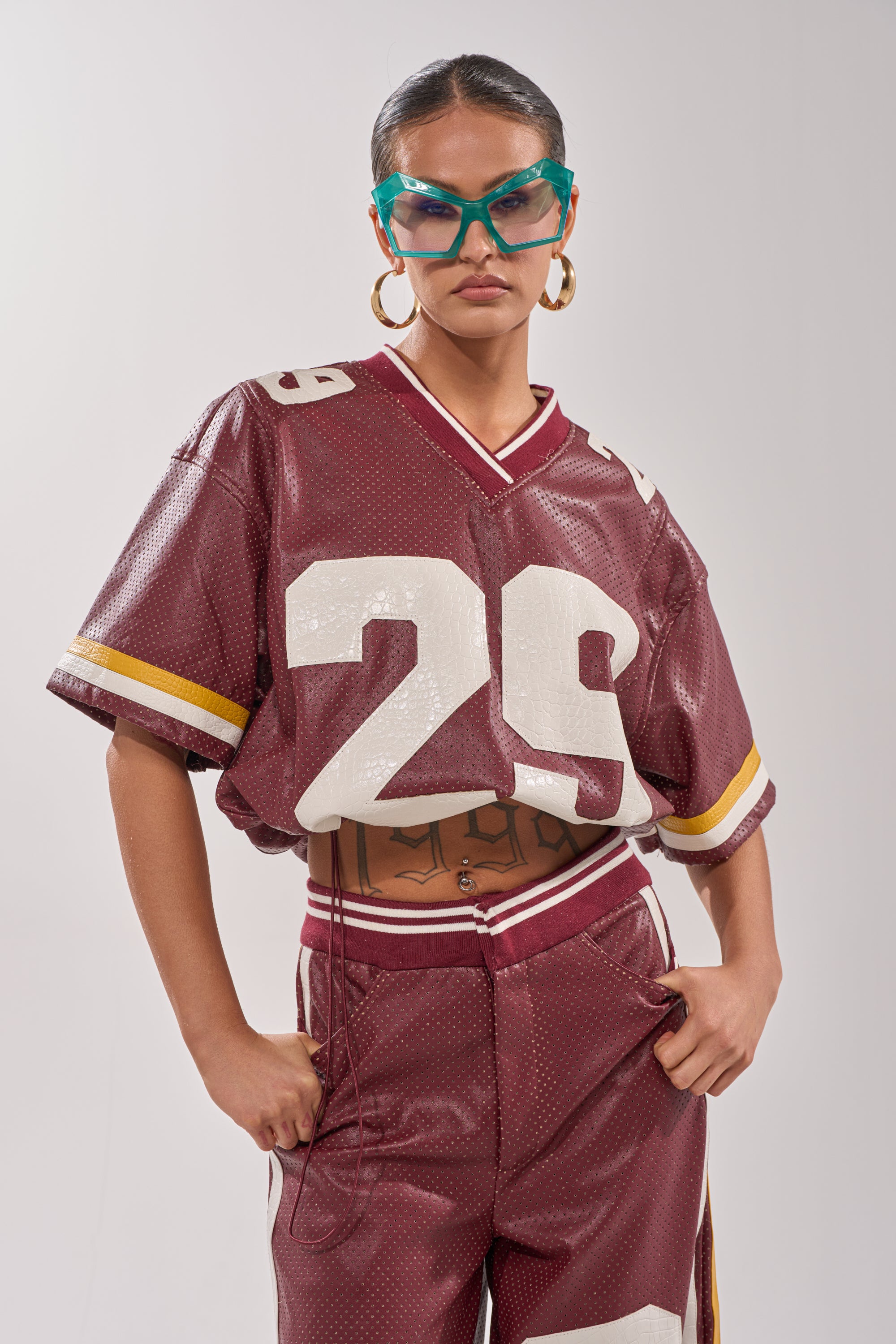 Wearing the LUCKY 29 JERSEY in burgundy with matching pants, bold teal sunglasses, and hoop earrings, a confident individual stands hands-on-hips against a plain background. 7