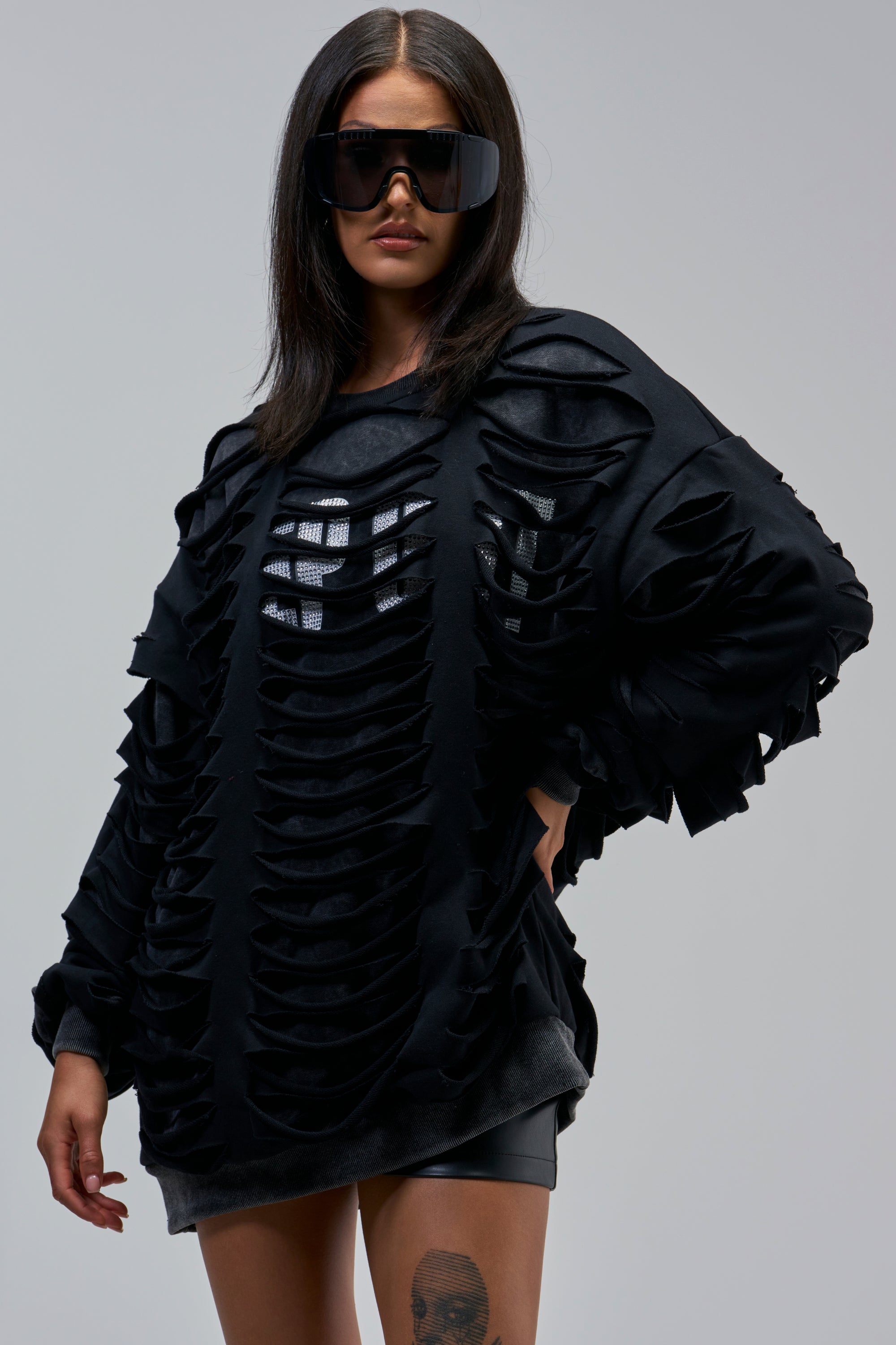 Woman wearing the EASILY DISTRESSED SWEATSHIRT with slashed cutouts over mesh, black shorts, statement sunglasses, and a thigh tattoo poses against a plain background for cool streetwear vibes.