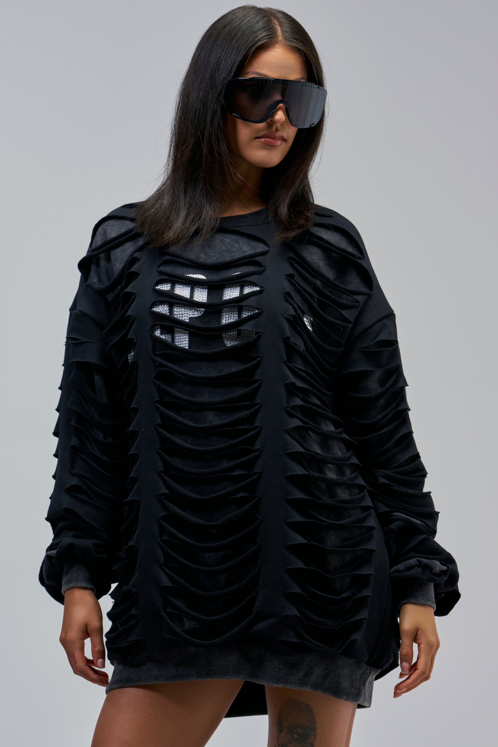 Woman wearing large black sunglasses and the EASILY DISTRESSED SWEATSHIRT with horizontal cutouts over a patterned shirt exudes bold streetwear style against a plain gray background.