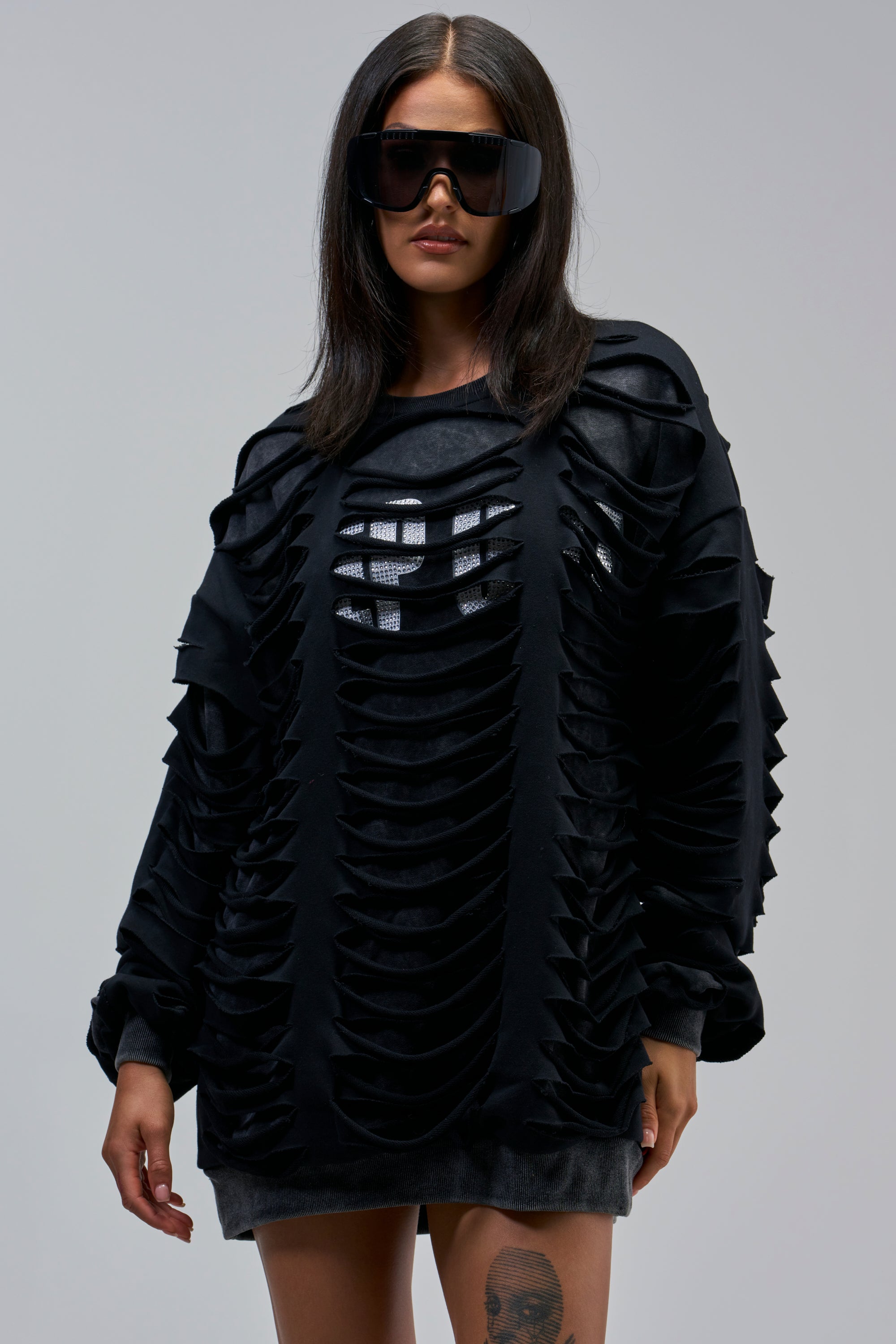 Woman wearing oversized black sunglasses and the EASILY DISTRESSED SWEATSHIRT, featuring layered cut-out designs. She stands against a plain background with straight dark hair and a tattoo on her right thigh.