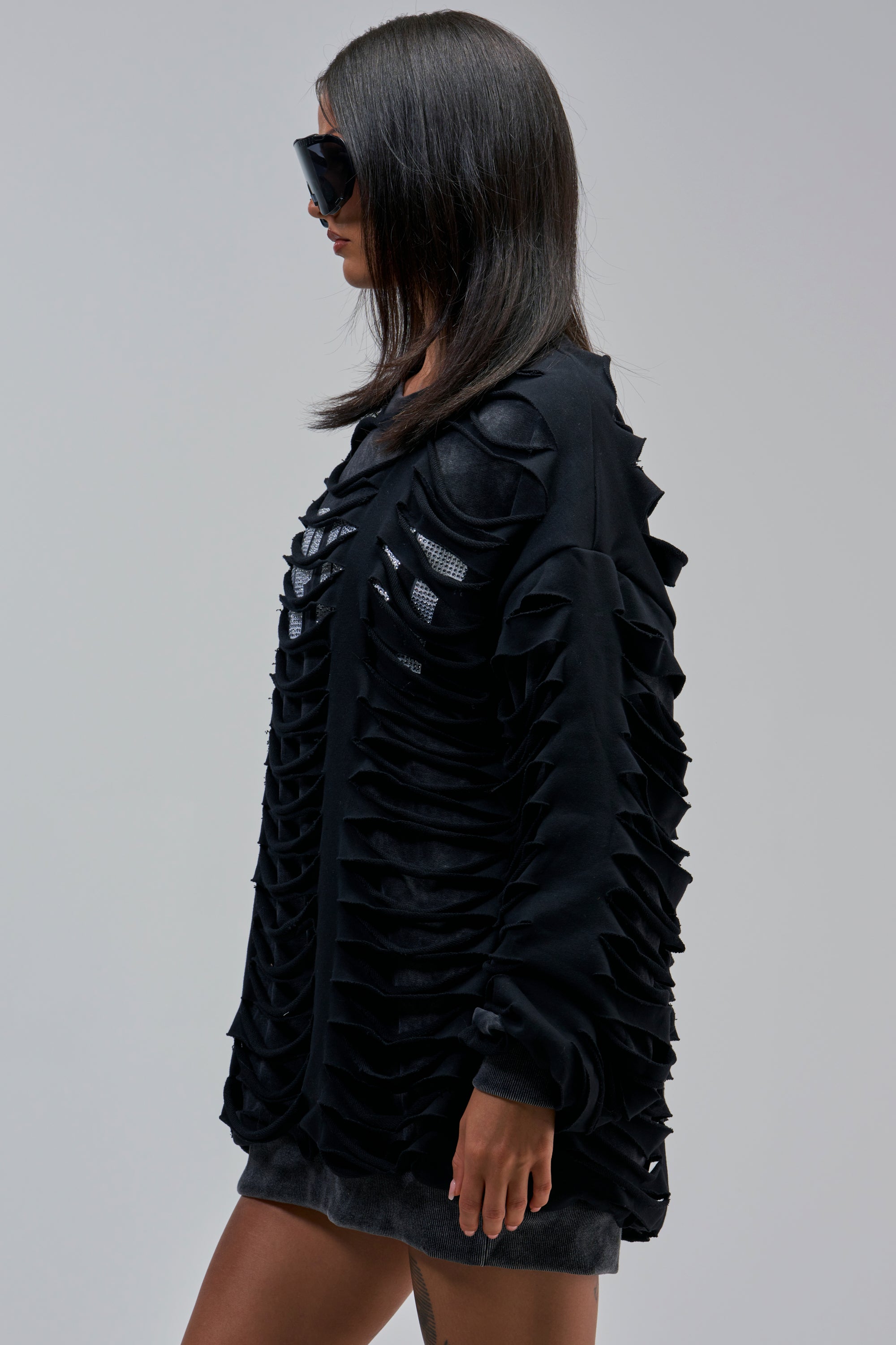 Woman in profile wears large black sunglasses and the EASILY DISTRESSED SWEATSHIRT with horizontal cutouts revealing a shimmery layer. She has straight, shoulder-length dark hair against a light background.