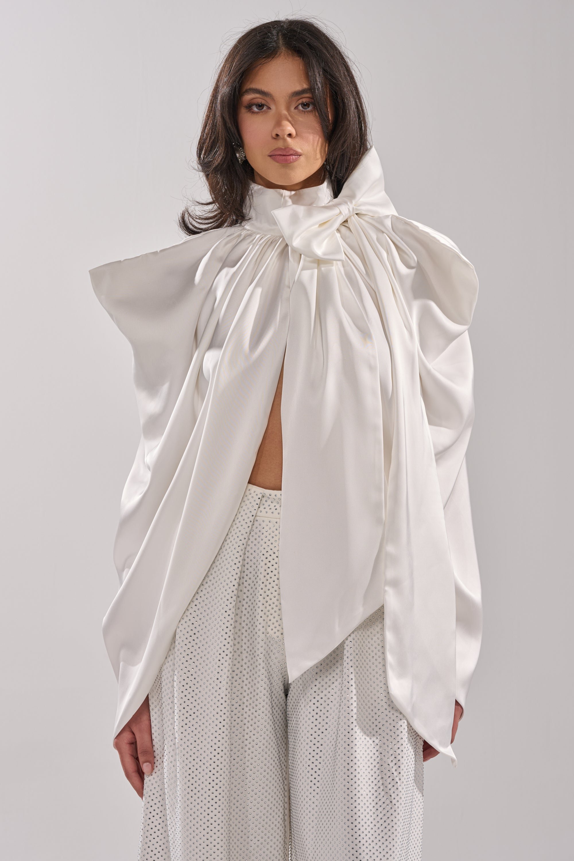 A person wears the WRAPPED IN LOVE BLOUSE, a dramatic white organza top with oversized sculptural ruffles and a bow at the neck, paired with white textured wide-leg pants, standing against a plain light background. 0