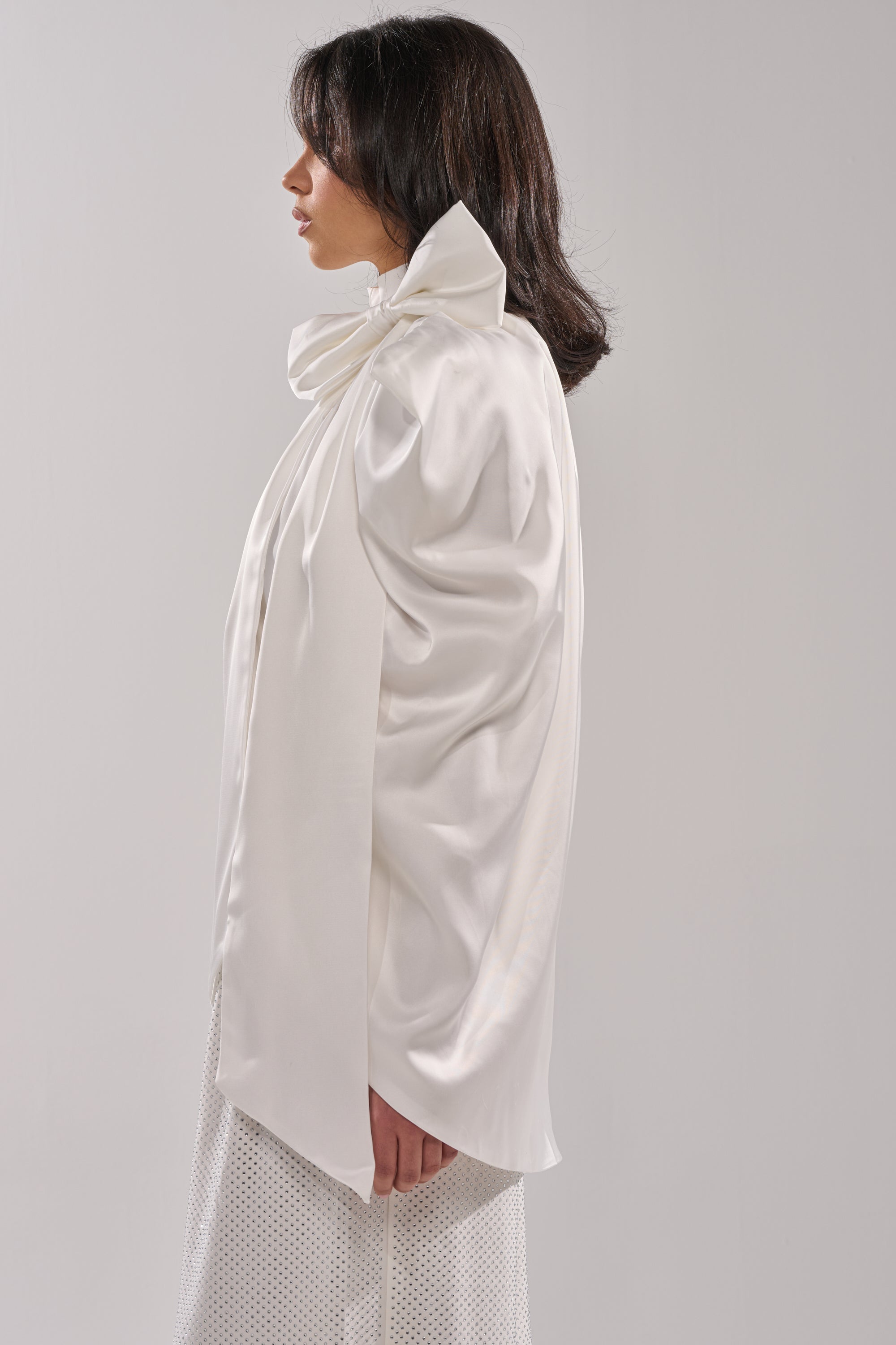 A woman in profile wears the WRAPPED IN LOVE BLOUSE, a high-fashion sheer top with an oversized sculptural neck bow and long, loose sleeves, styled with a light textured skirt and dark shoulder-length hair against a plain background. 1