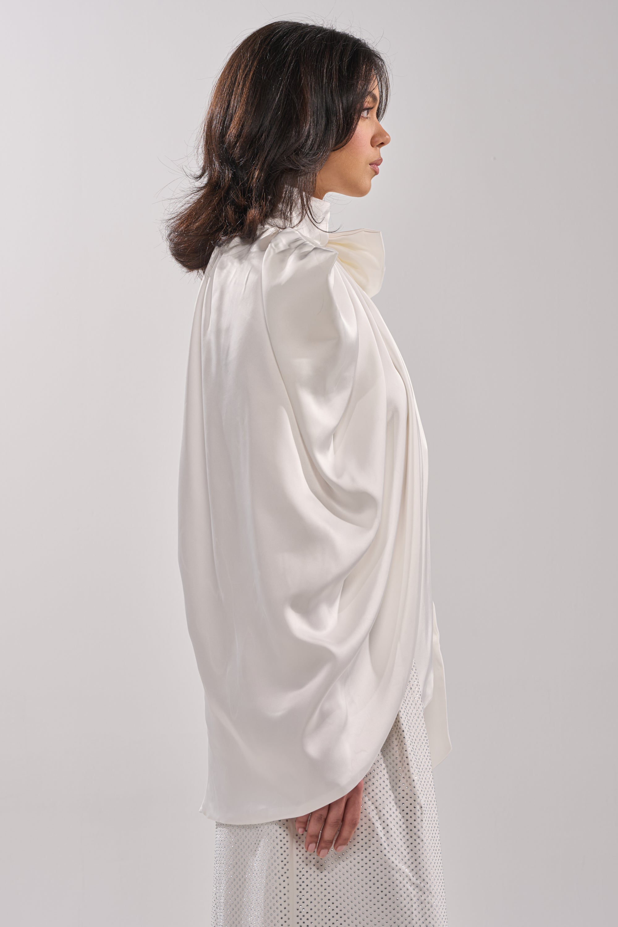 A woman with dark hair stands in profile wearing the shiny white WRAPPED IN LOVE BLOUSE and a white textured skirt against a plain light background. 2
