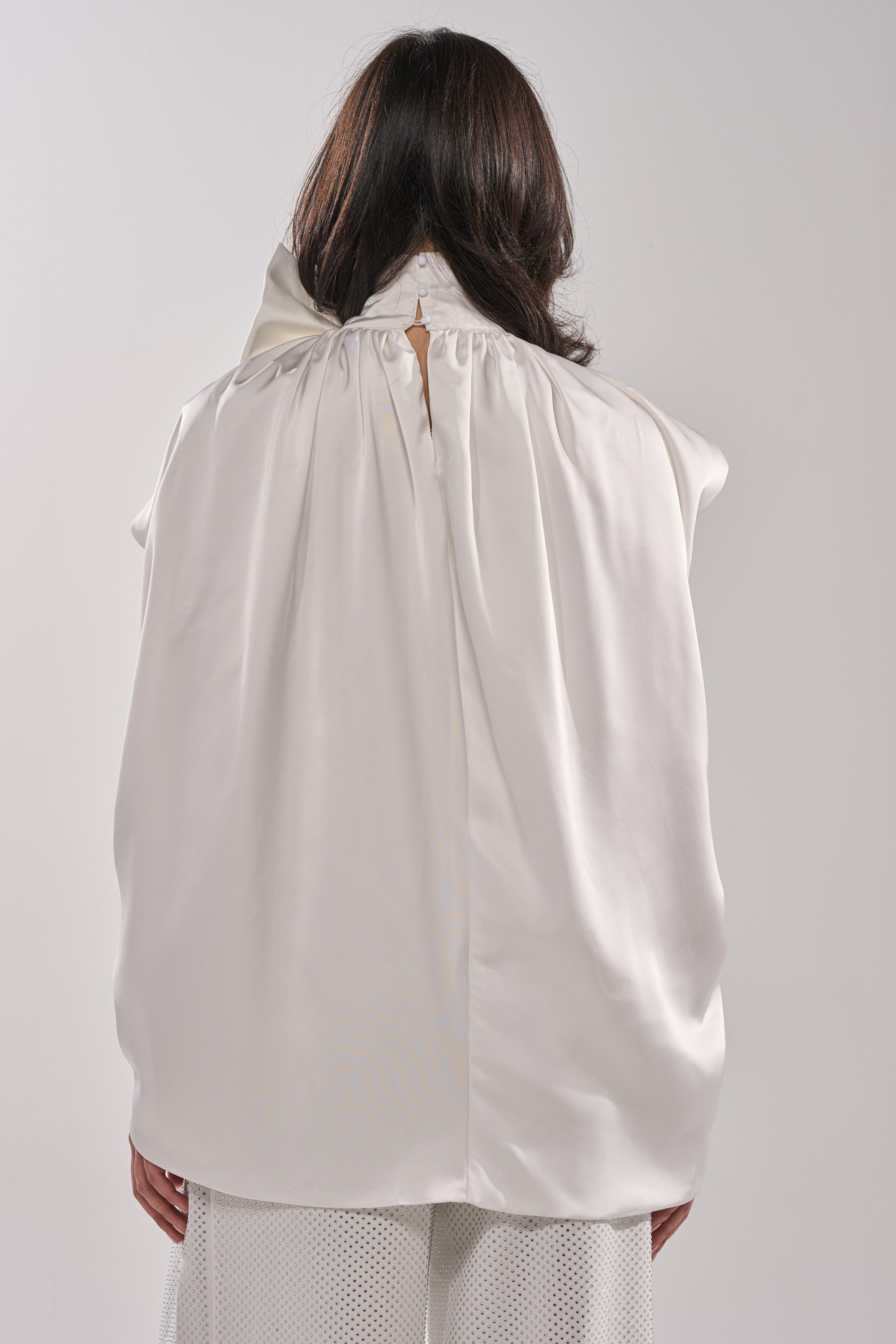 A person with shoulder-length dark hair is seen from the back, wearing the WRAPPED IN LOVE BLOUSE—a loose-fitting white organza top with a gathered neckline and wide sleeves—paired with white textured pants against a plain light background. 3