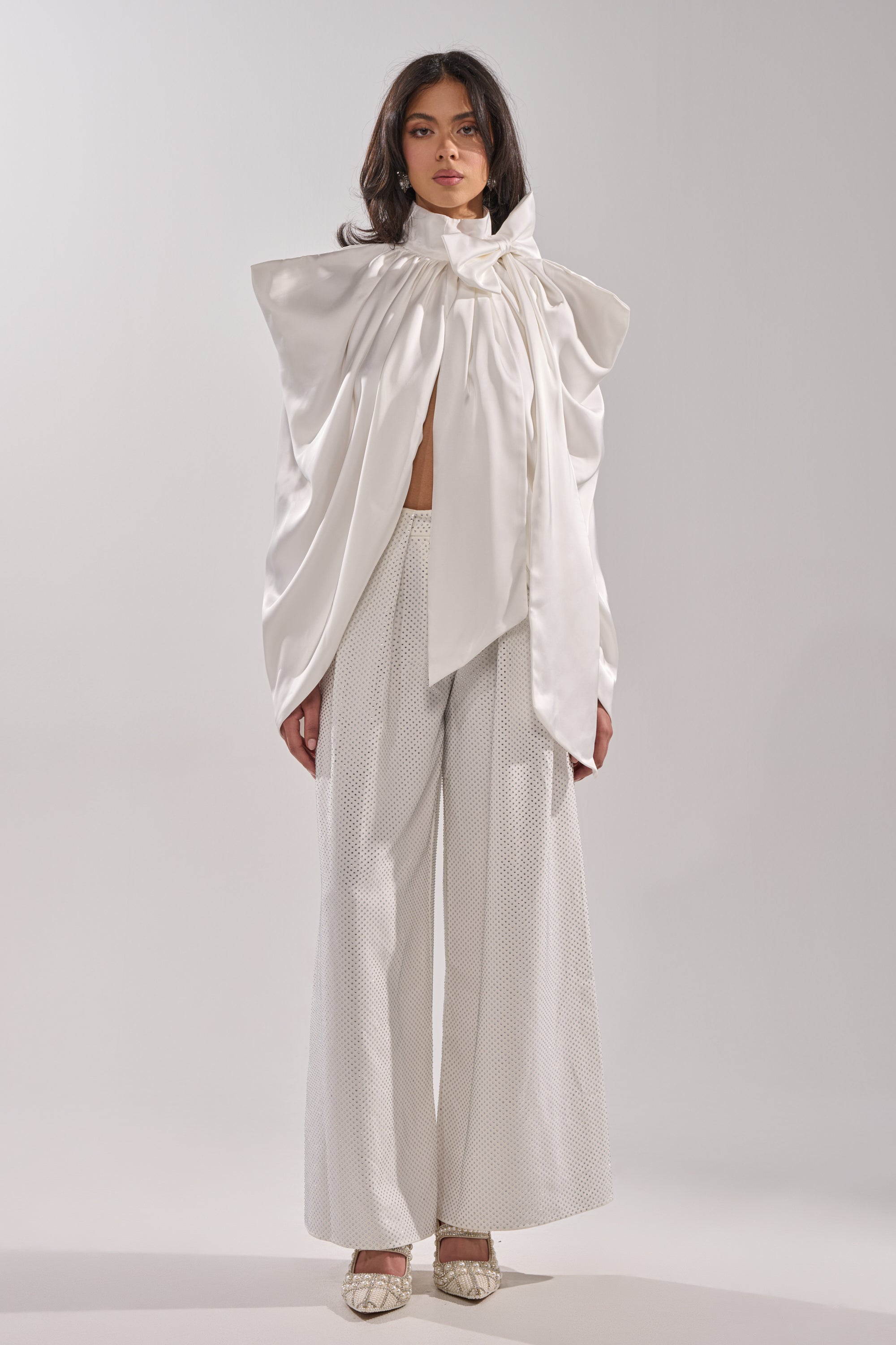 A woman faces forward in the WRAPPED IN LOVE BLOUSE, featuring a dramatic sculptural bow and oversized white satin drapes, paired with textured wide-leg trousers and pointed-toe shoes against a plain light background. 4