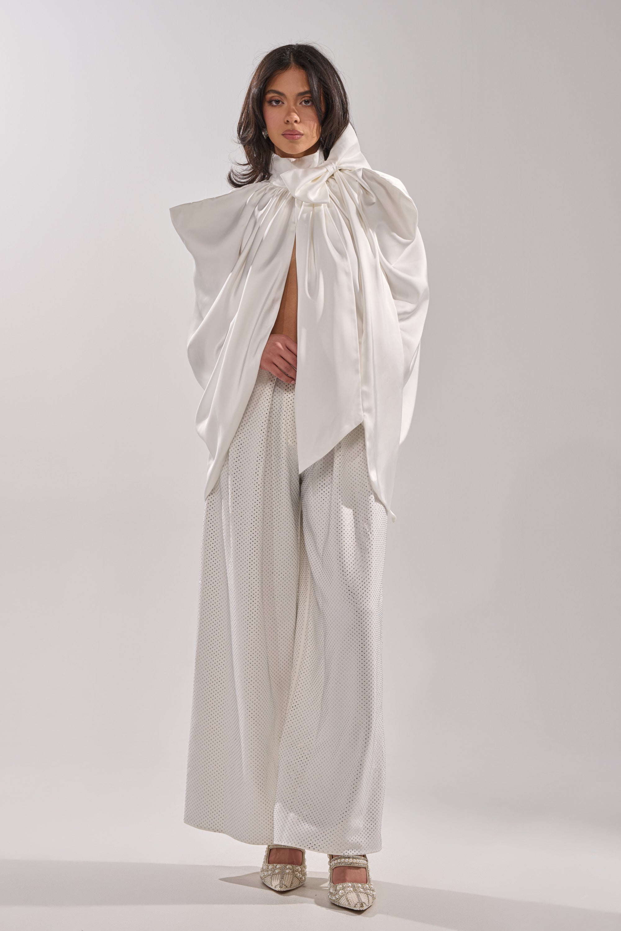 A woman stands against a plain background wearing the WRAPPED IN LOVE BLOUSE in voluminous white satin with draped sleeves and wide-leg textured white pants, finished with pointed heels. 5